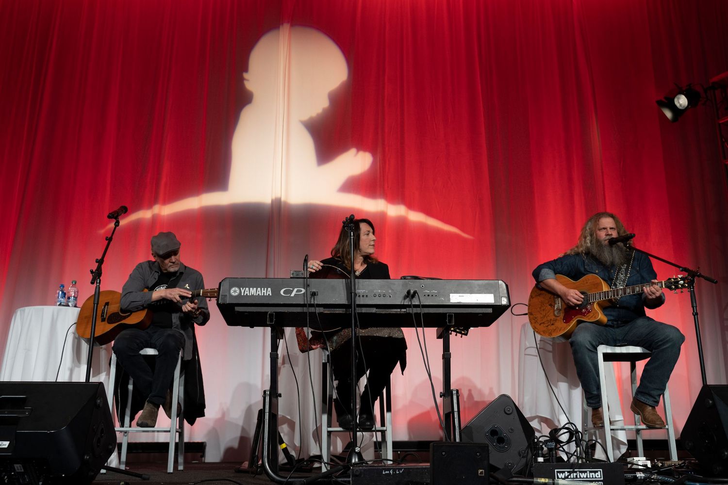 Jamey Johnson performing acoustic music at a St. Jude Children’s Research Hospital charity pasta dinner event on a red-curtained stage.