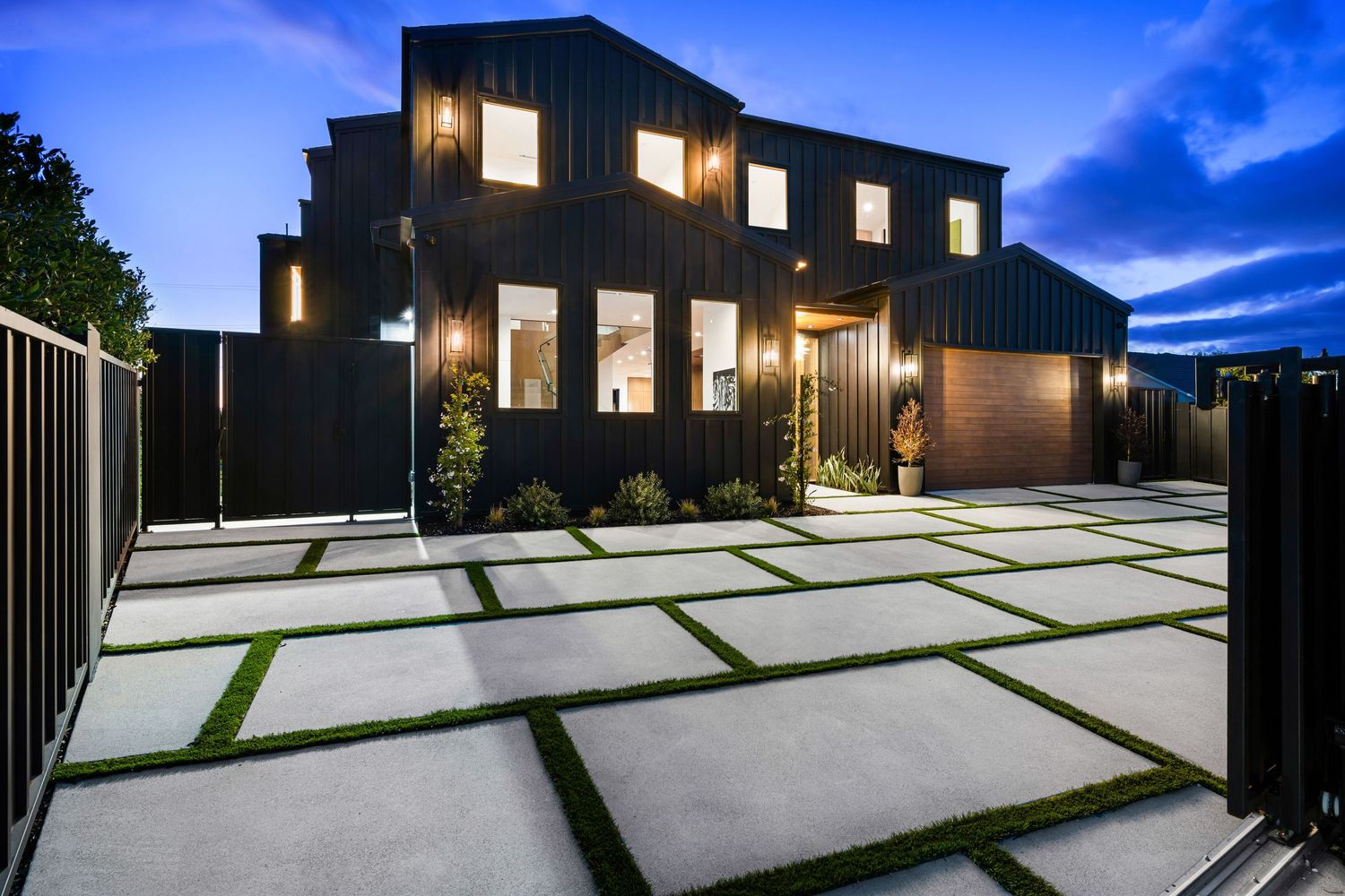 Modern black home with large concrete paver driveway and green grass accents illuminated at dusk.