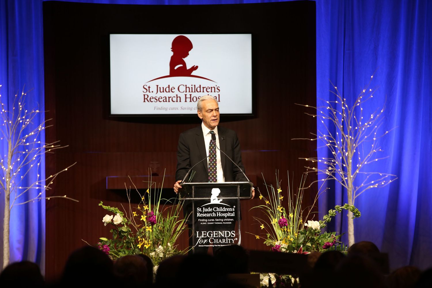 A speaker presents on stage at a St. Jude Children's Research Hospital event with blue curtains and decorative branches.