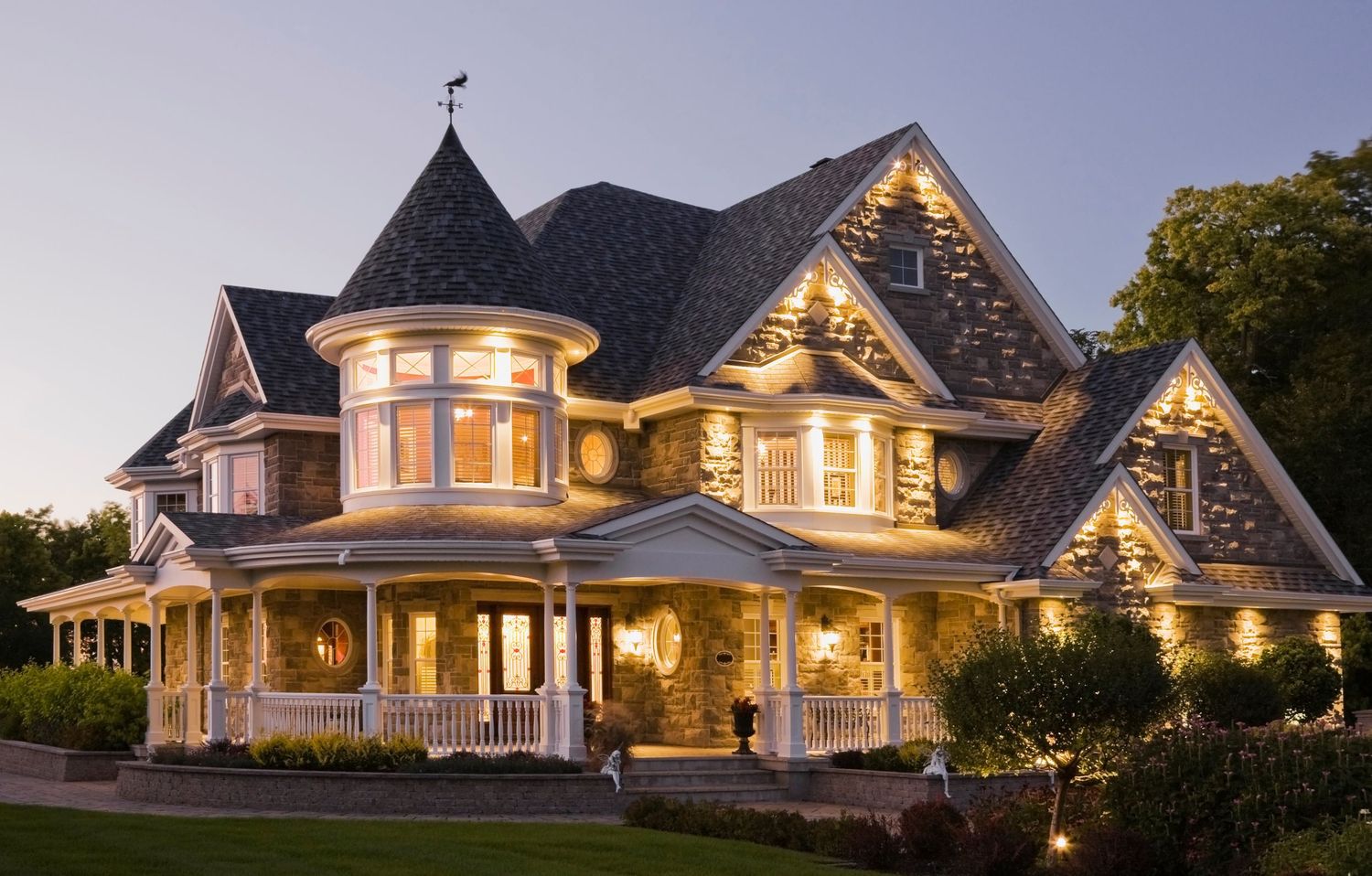 Stunning Victorian mansion with wrap-around porch, turret, and warm glowing lights at dusk.