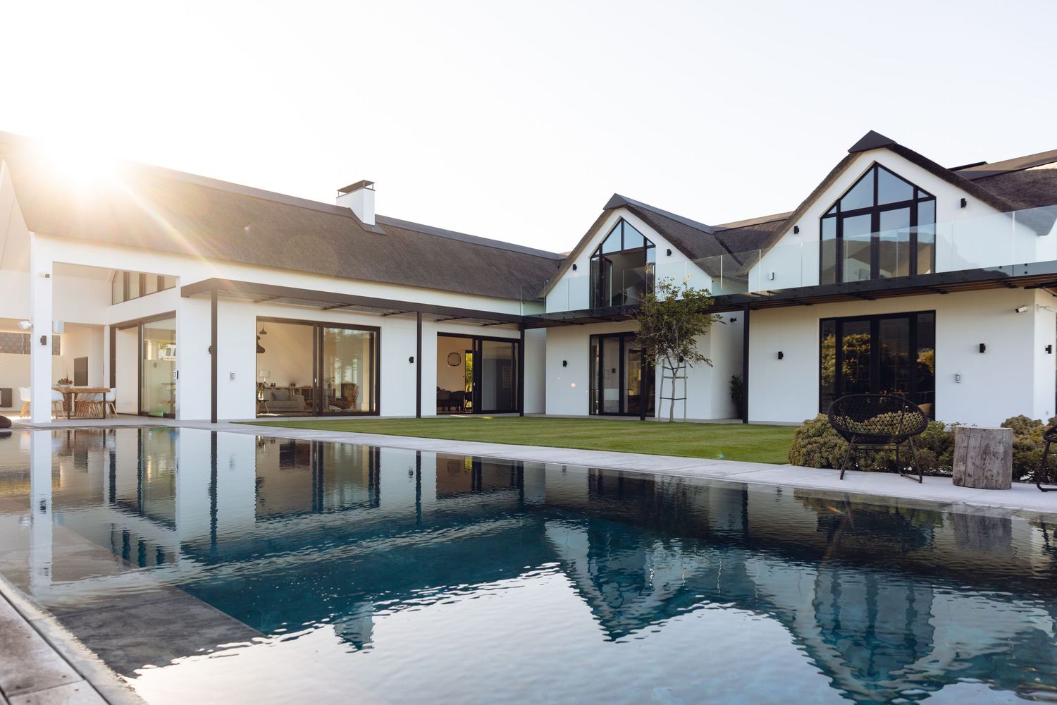 Modern white farmhouse with black-framed windows and a sleek rectangular pool reflecting sunlight in a manicured backyard.