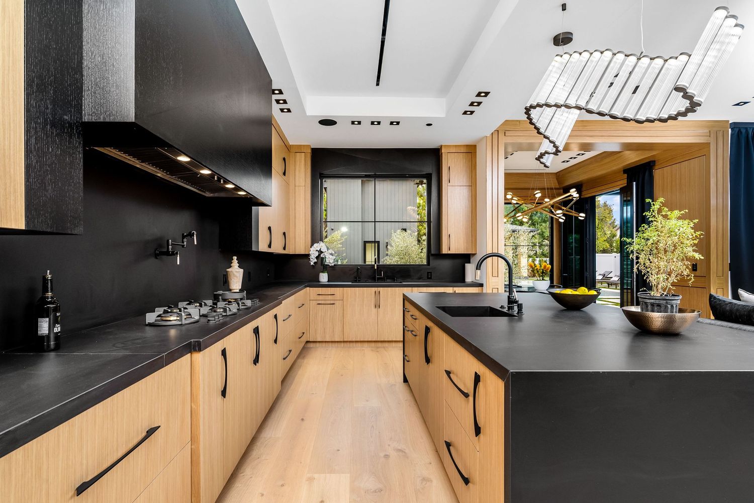 Modern kitchen with black countertops, light wood cabinets, gas cooktop, and elegant pendant lighting.