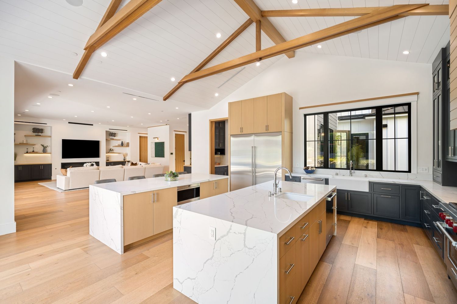 Modern open-concept kitchen with white quartz island, light wood cabinets, vaulted beam ceilings, and hardwood floors.