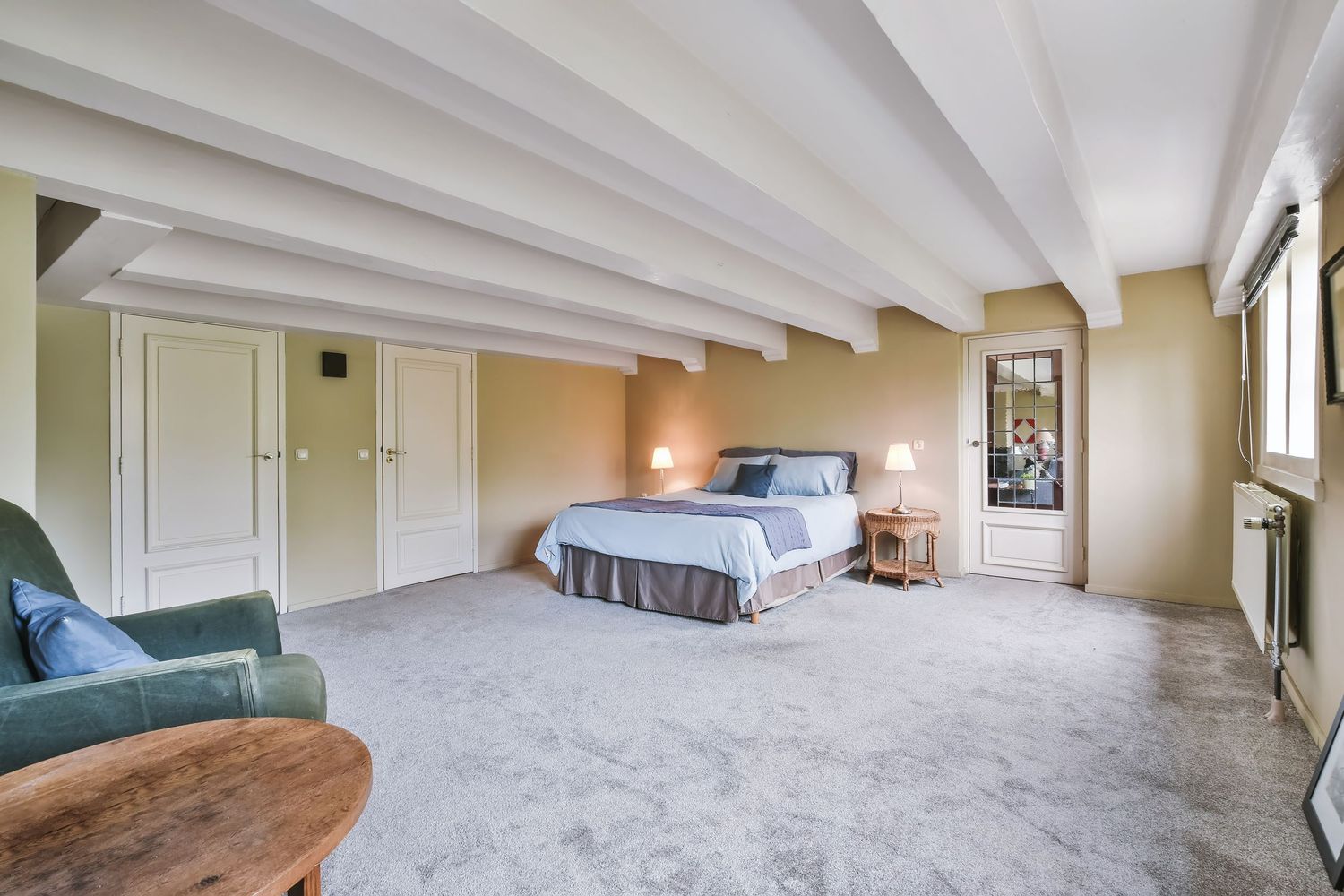 Spacious bedroom with exposed beam ceiling, plush carpet, a queen bed, green armchair, and white built-in closets.