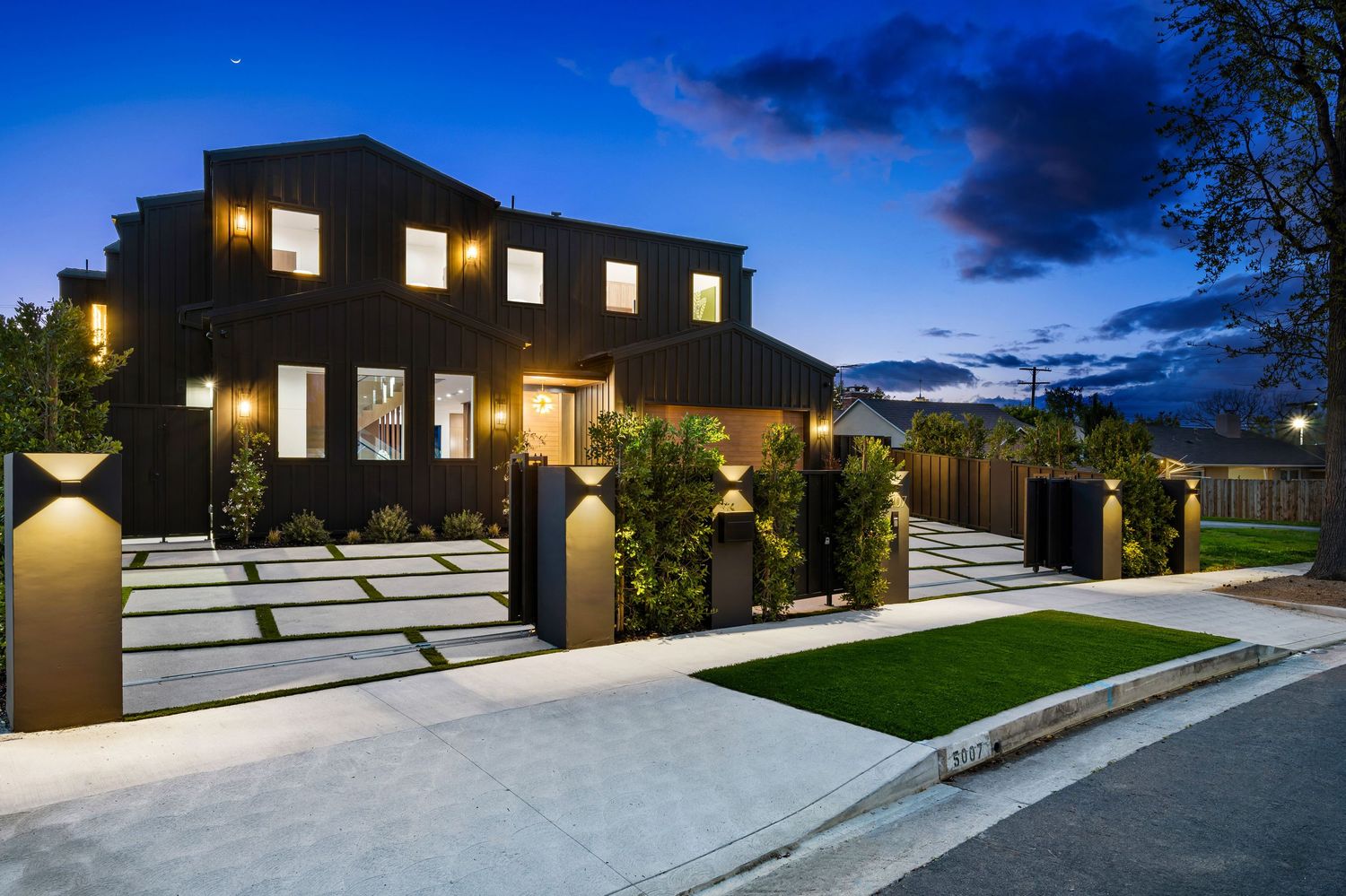 Modern two-story dark exterior home with illuminated landscaping, concrete driveway, and manicured lawn at twilight.