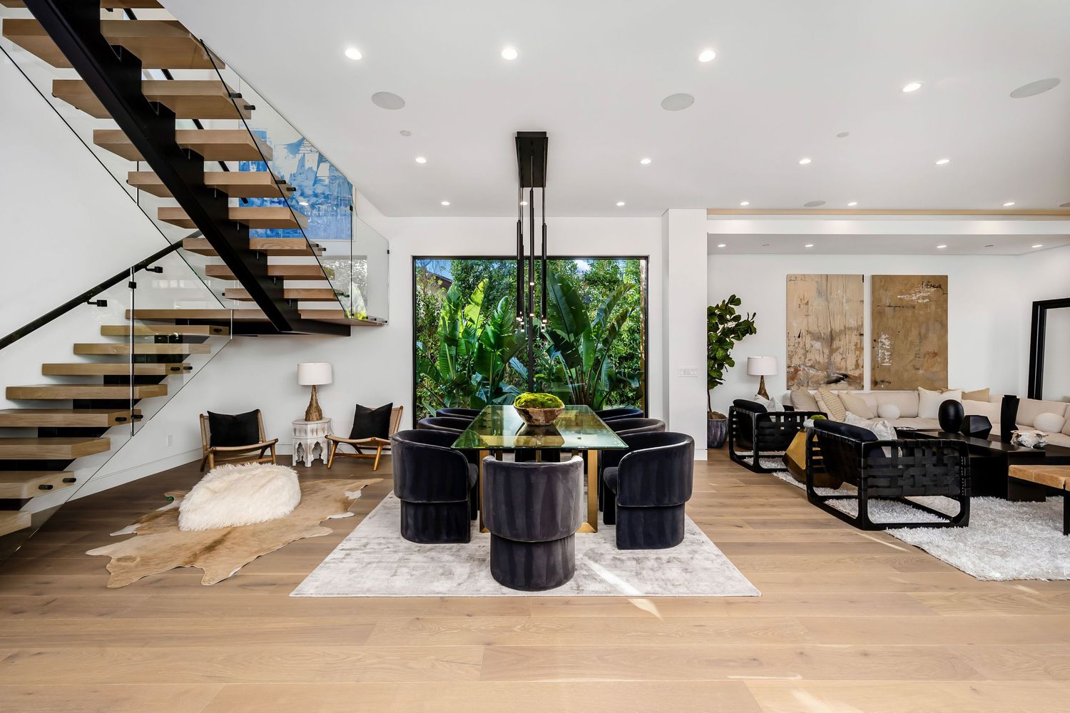 Modern open-plan dining room with black chairs, glass table, floating staircase, and tropical garden view through floor-to-ceiling windows.