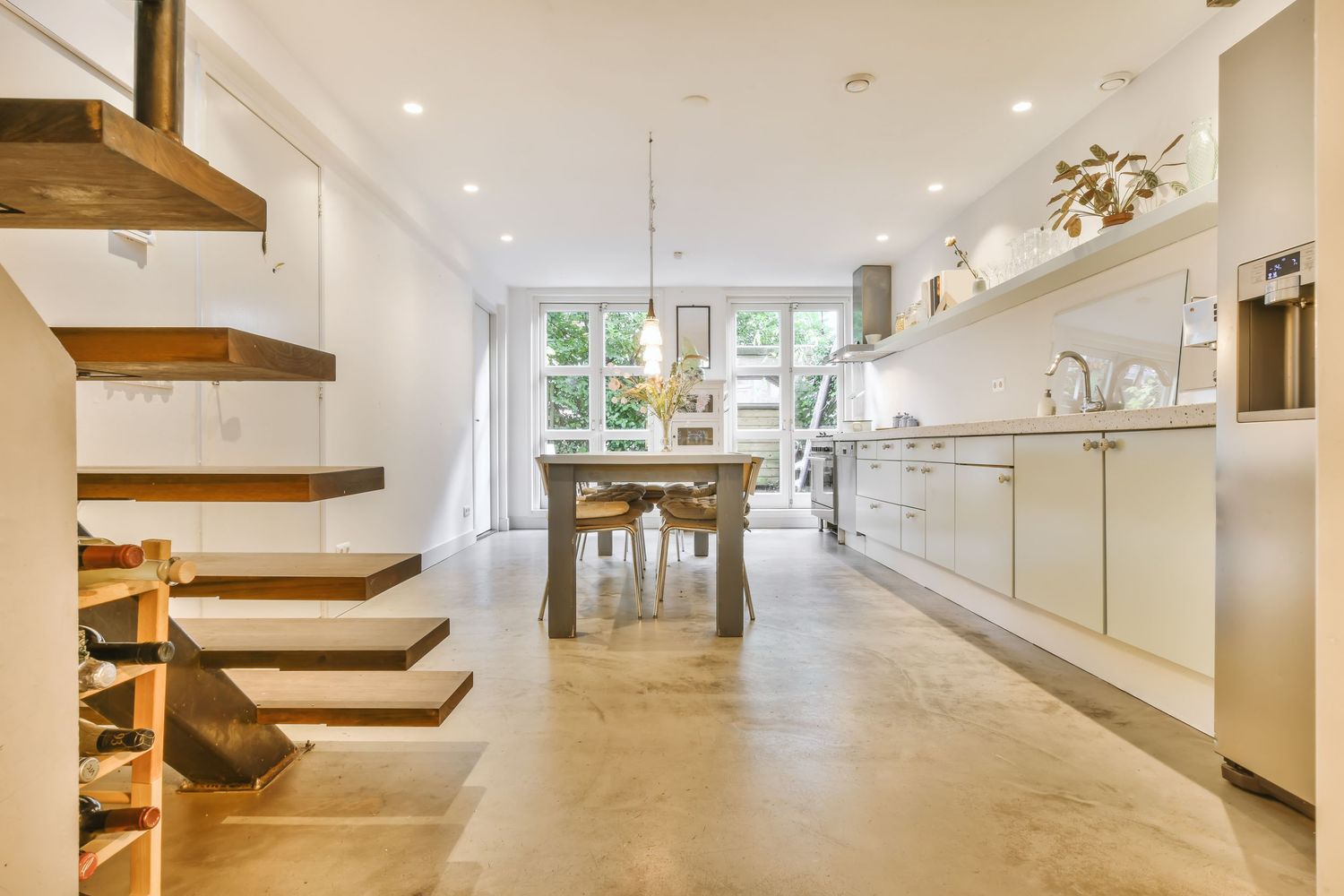 Modern open-plan kitchen and dining area with floating staircase, concrete floors, white cabinetry, and natural light from large windows.