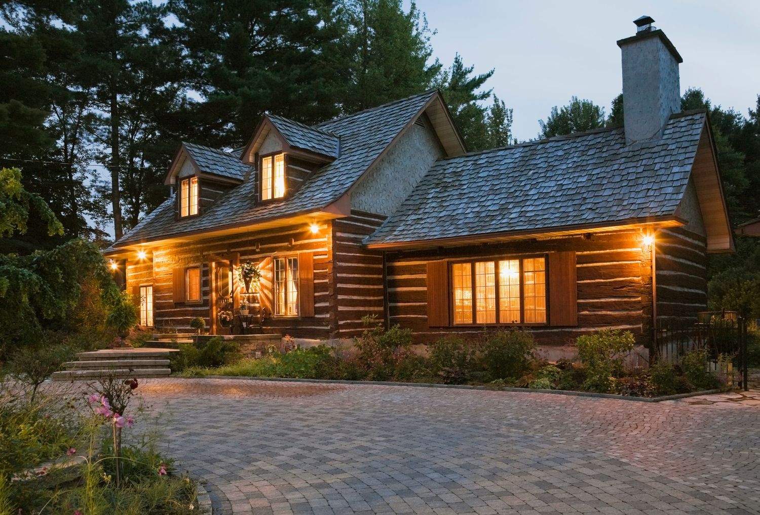 Rustic log cabin home with warm glowing windows, stone chimney, cobblestone driveway, and lush trees at dusk.