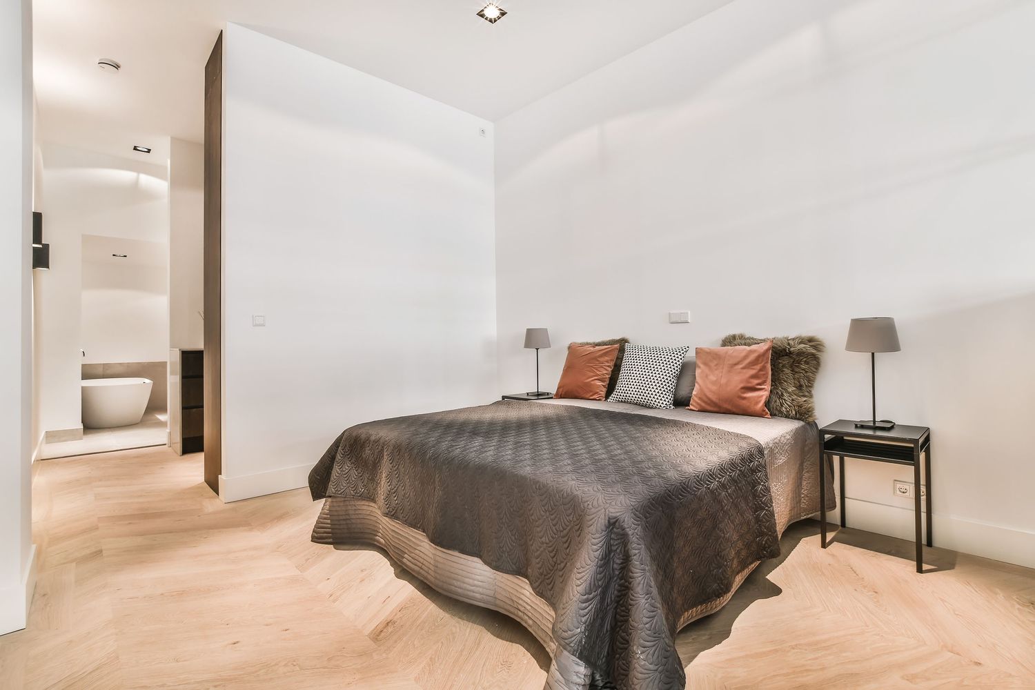 Modern bedroom with gray quilted bedspread, terracotta pillows, bedside lamps, and ensuite bathroom on light wood floors.