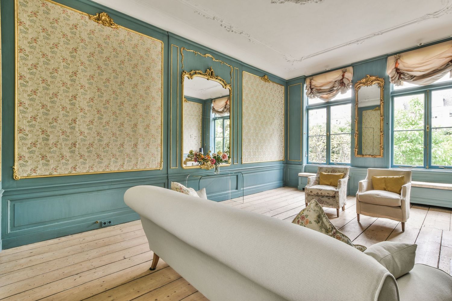 Elegant French-style living room with teal walls, gold mirrors, floral wallpaper, hardwood floors, and vintage armchairs.