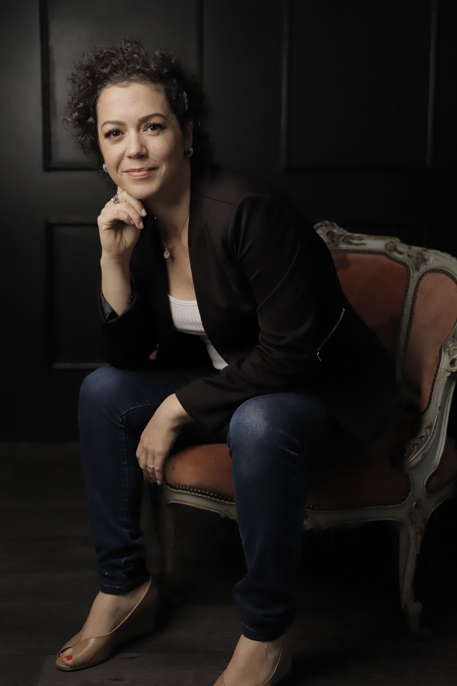 Woman in black blazer and jeans sits thoughtfully in vintage chair against dark background.