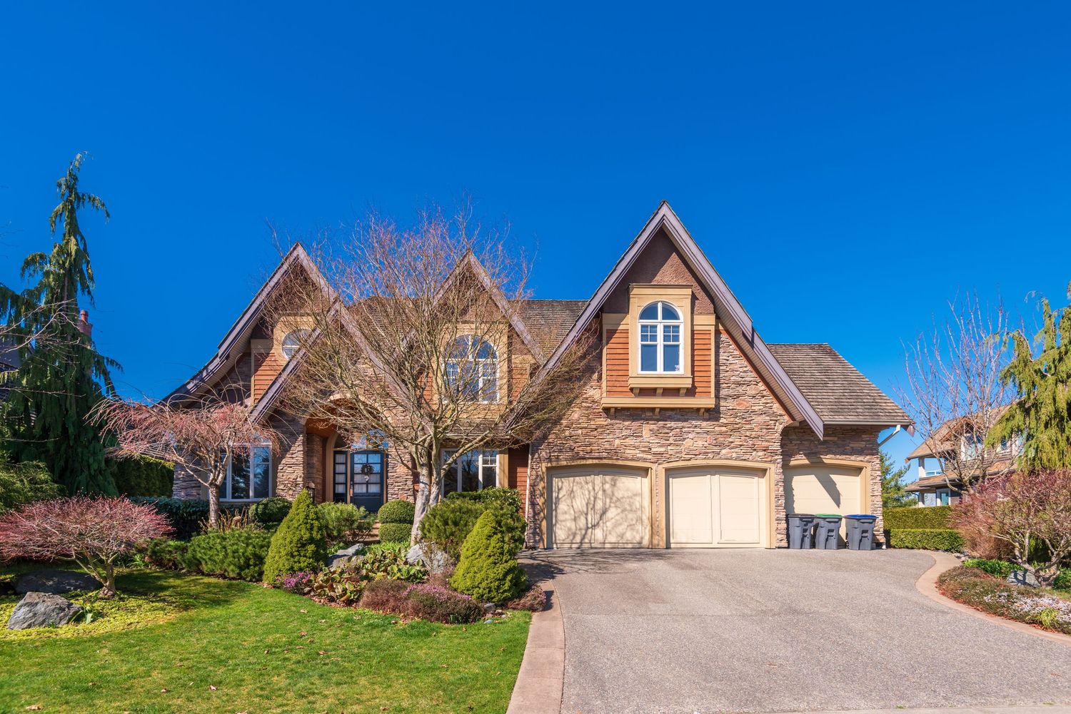 Elegant brick Tudor-style home with manicured landscaping, arched windows, double garage, and a curved driveway under a clear blue sky.