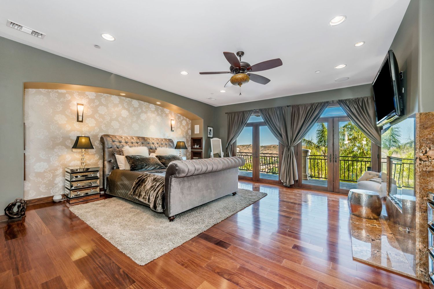 Luxurious master bedroom with hardwood floors, tufted bed, accent wall, ceiling fan, and French doors opening to a scenic balcony.