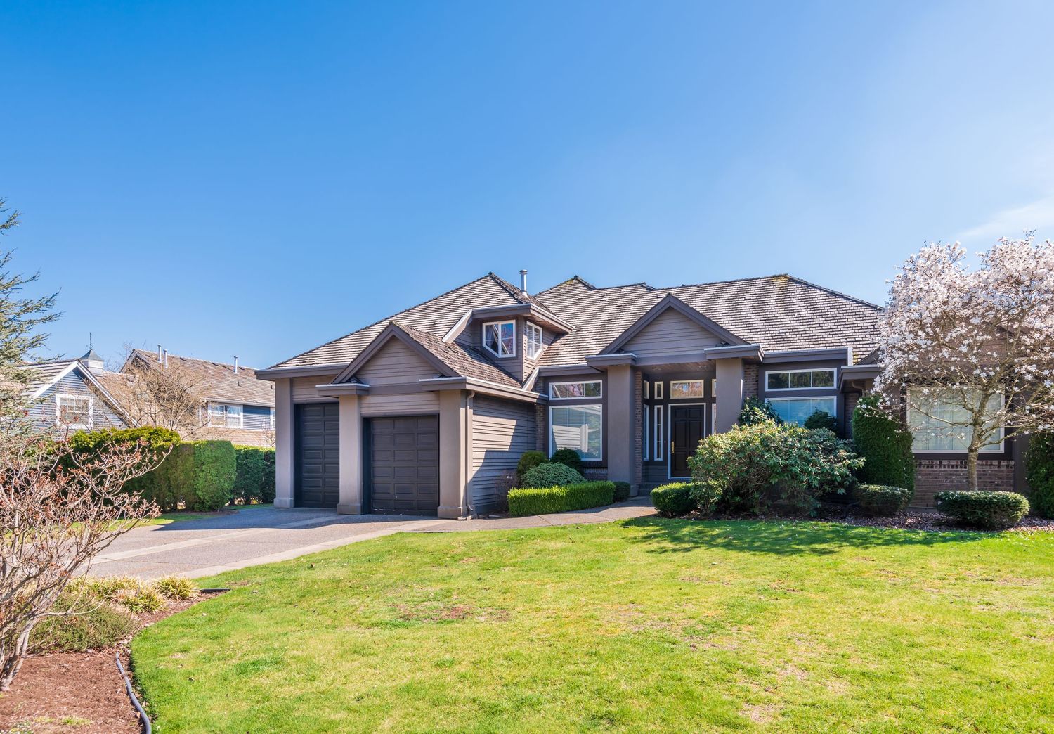 Single-story suburban home with double garage, manicured lawn, and blooming trees under a clear blue sky.