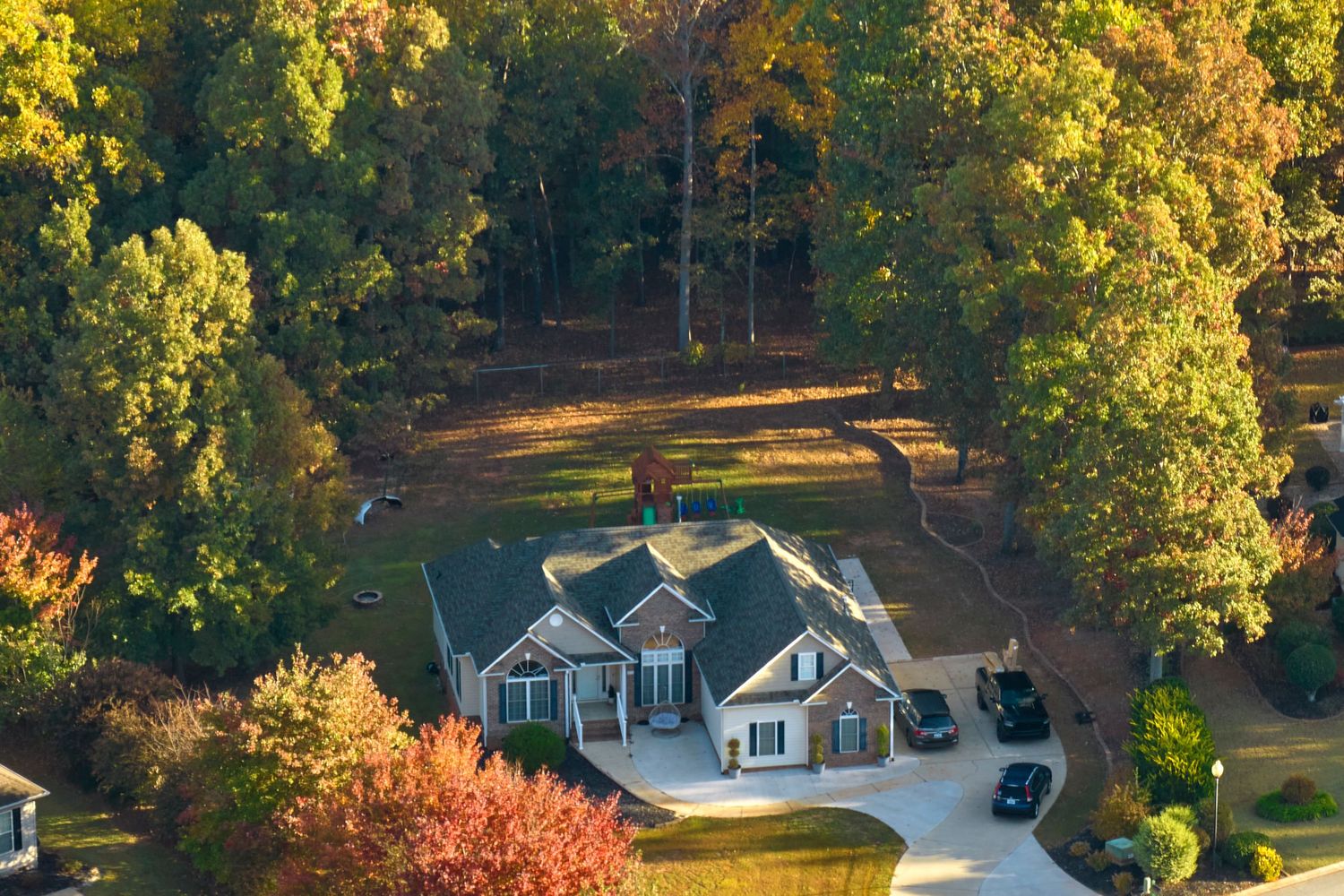 Aerial view of a large suburban home surrounded by autumn trees with a circular driveway and open backyard.
