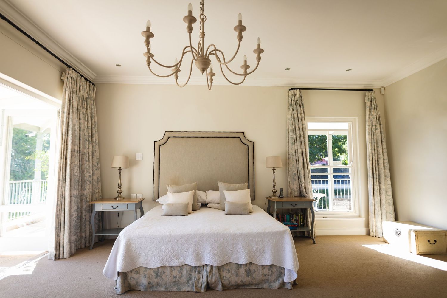 Elegant bedroom with chandelier, upholstered headboard, white bedding, floral curtains, and French doors.