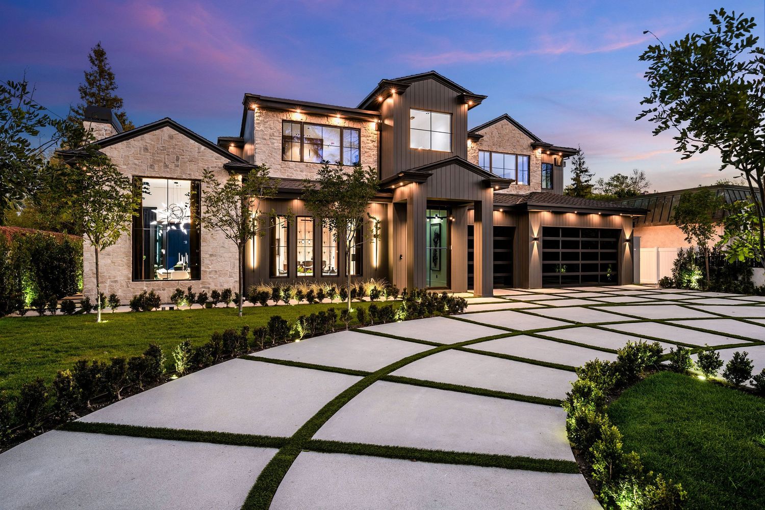 Luxury modern stone mansion at dusk with illuminated geometric driveway, manicured lawn, and elegant architectural details.
