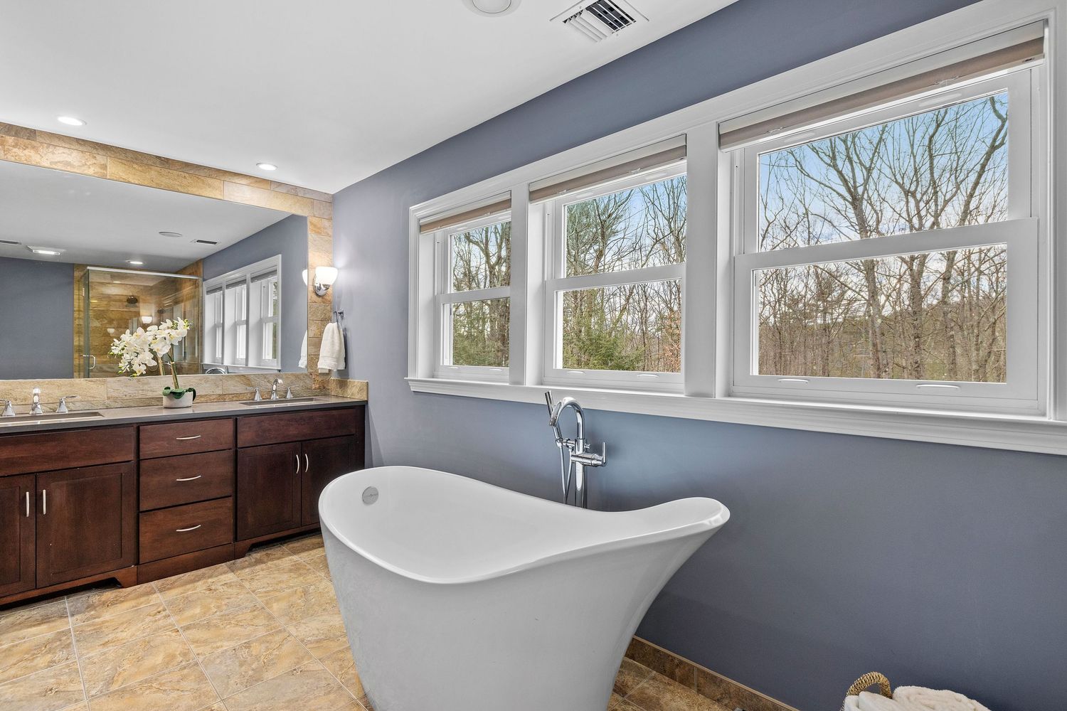 Elegant master bathroom featuring freestanding soaking tub, double vanity, and large windows with wooded views.