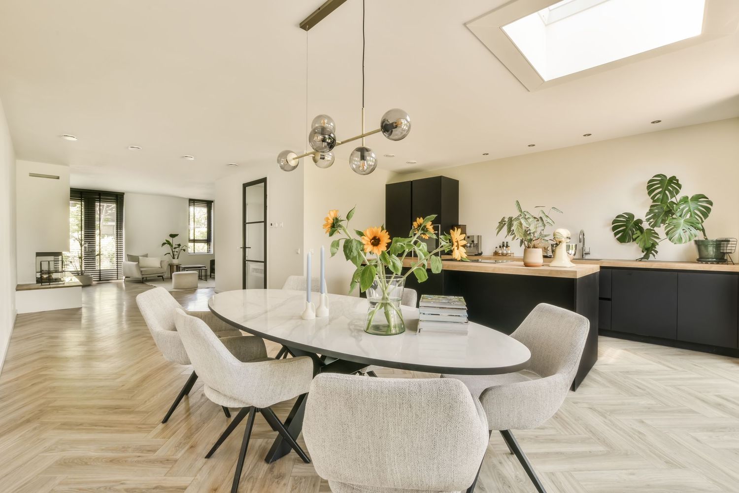 Modern dining room with oval marble table, gray chairs, pendant light, herringbone floors, and sunflowers centerpiece.