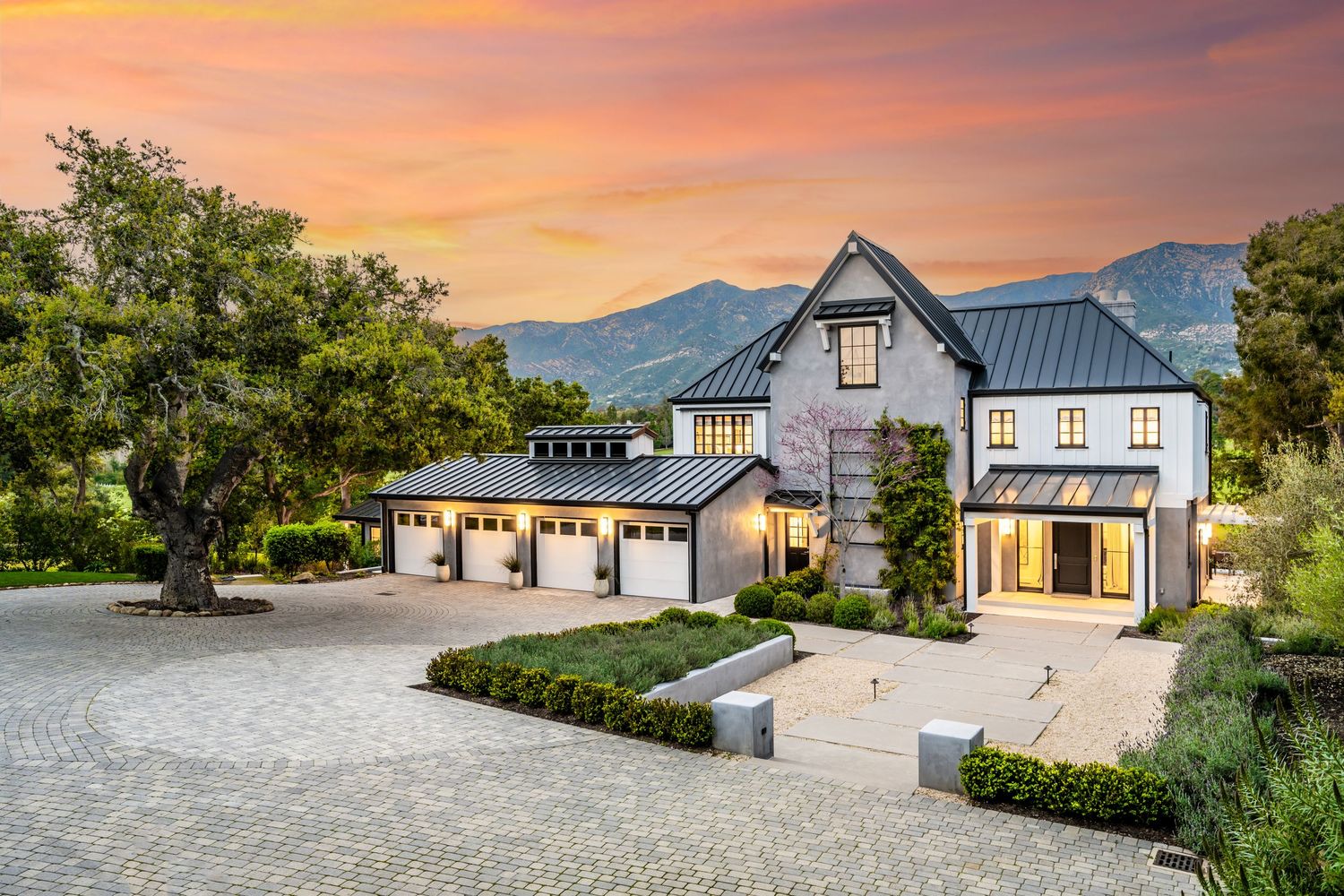 Luxury estate home with metal roof, three-car garage, cobblestone driveway, and mountain backdrop at sunset.