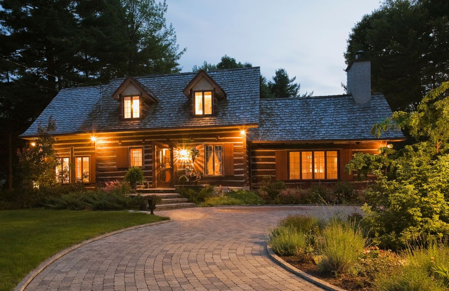 Charming log cabin home exterior illuminated at dusk with warm lights, stone pathway, and lush landscaping.
