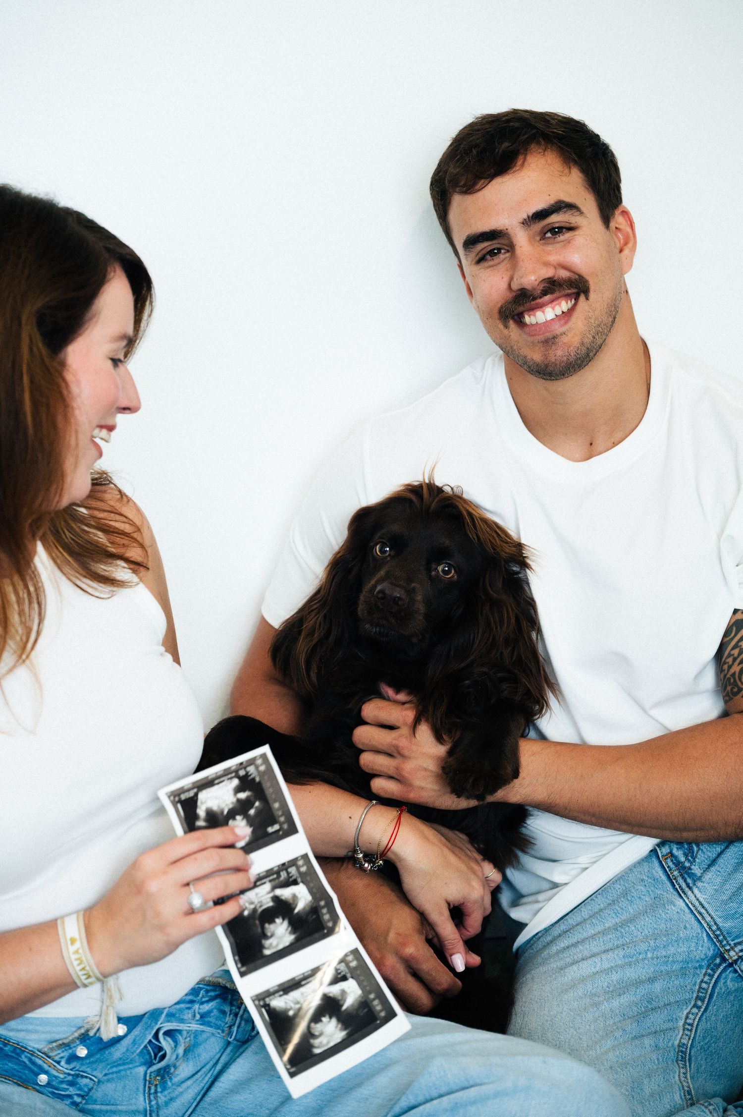 Smiling couple in white shirts sit with a black cocker spaniel while holding ultrasound photos in a pregnancy announcement.