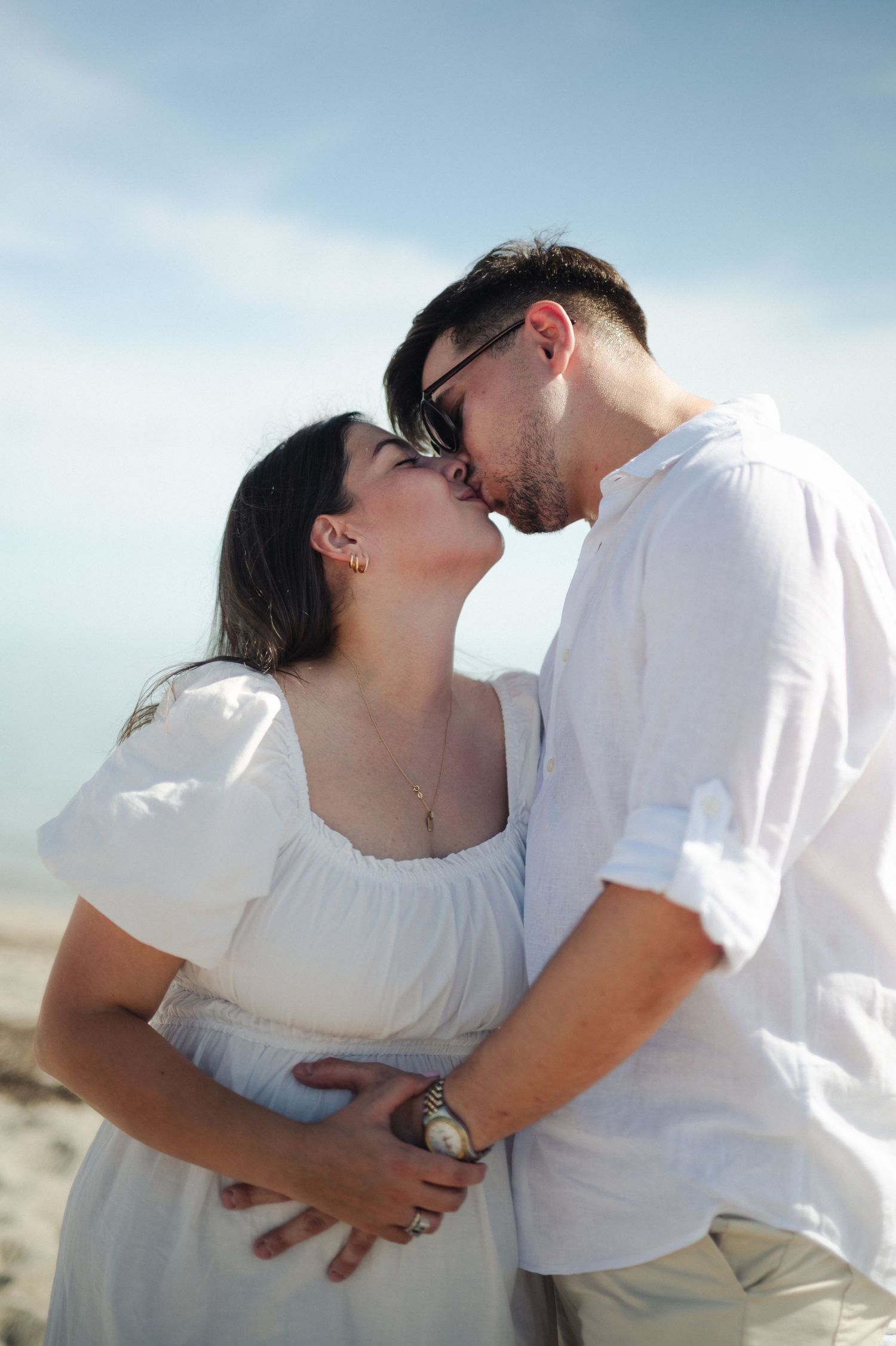 Couple in white linen outfits share a kiss on the beach as the man gently holds her pregnant belly.