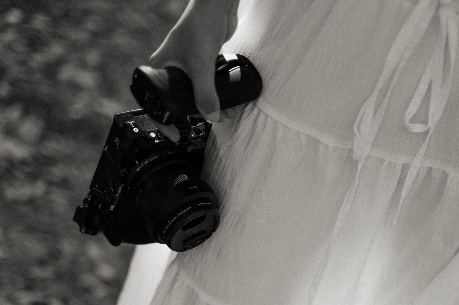Black and white photo of a woman in a flowing white dress holding a vintage camera at her side.