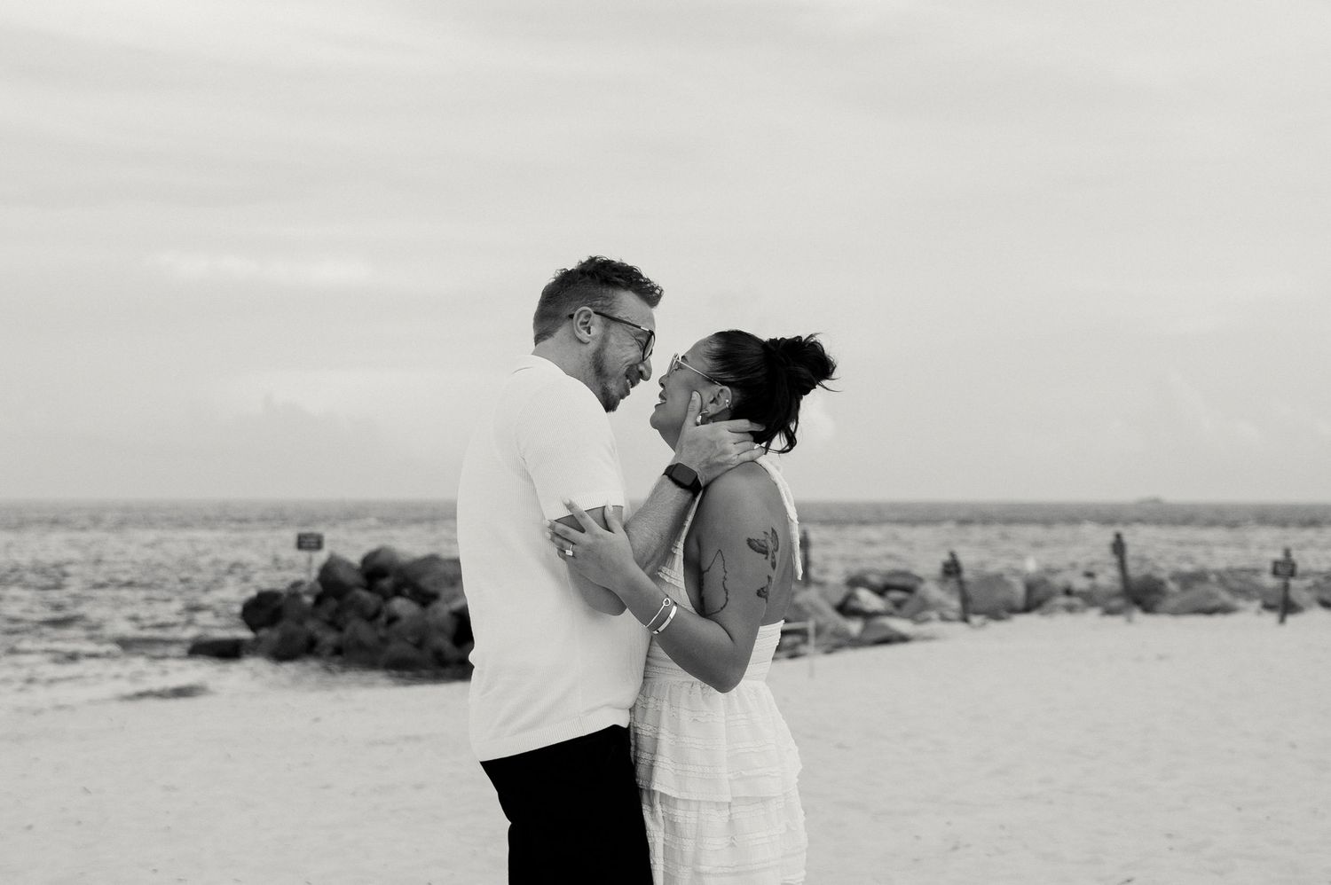 Black and white photo of a couple embracing and kissing on a beach near the ocean.