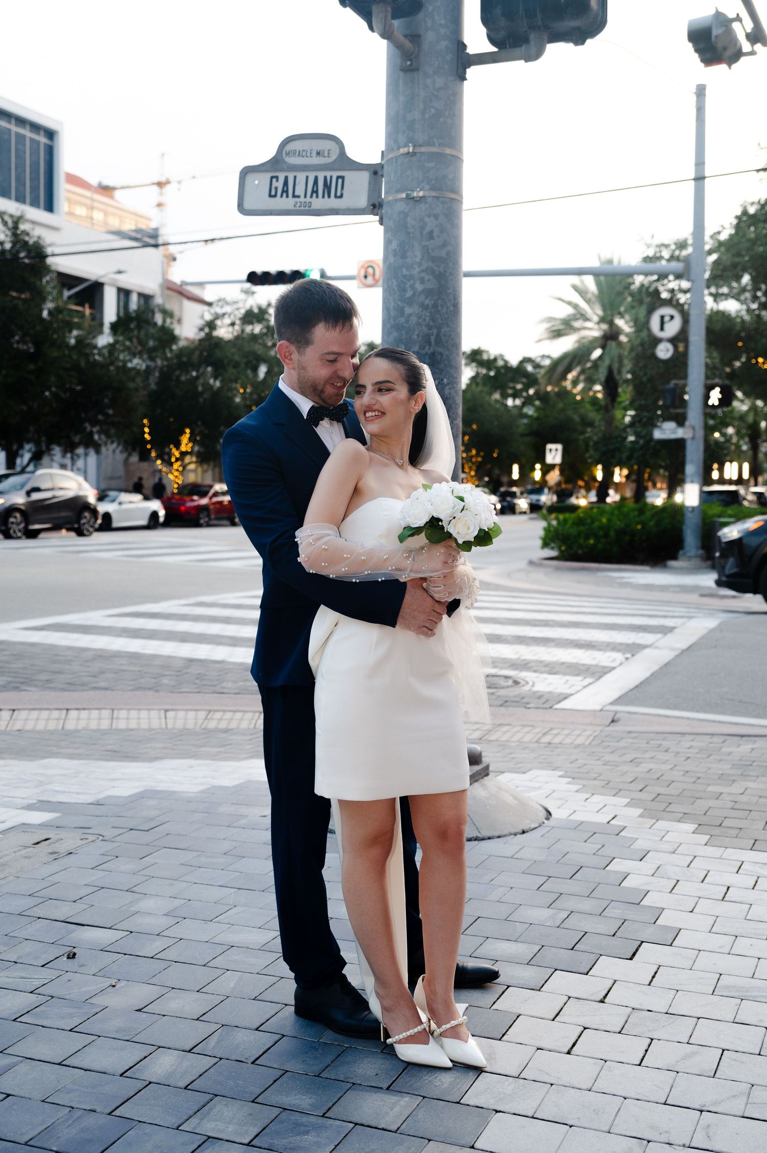 Bride holding a white bouquet and wearing white heels is embraced by her groom beside a Galiano street sign at their city wedding.