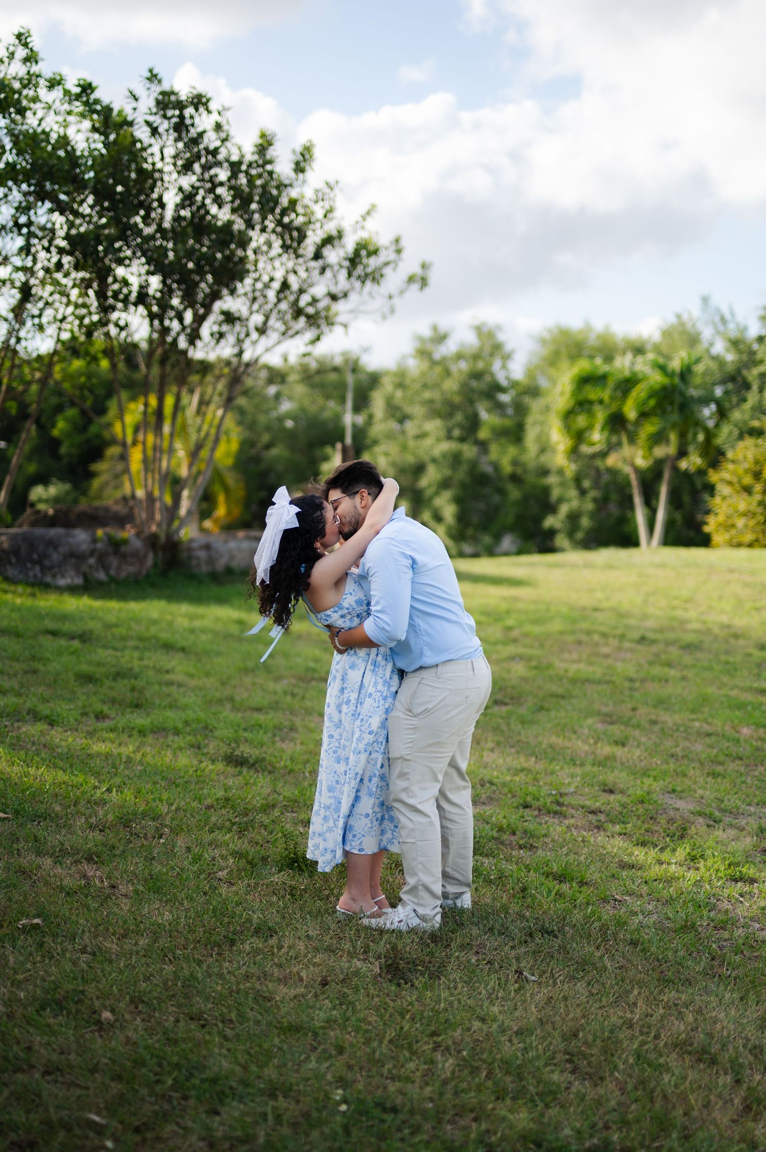 Couple in light blue outfits embracing and kissing on a green lawn surrounded by trees and palm trees.