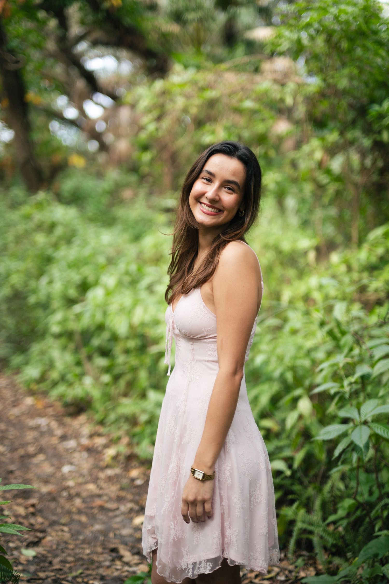 Smiling young woman in a light pink lace dress poses on a shaded nature trail surrounded by green foliage.
