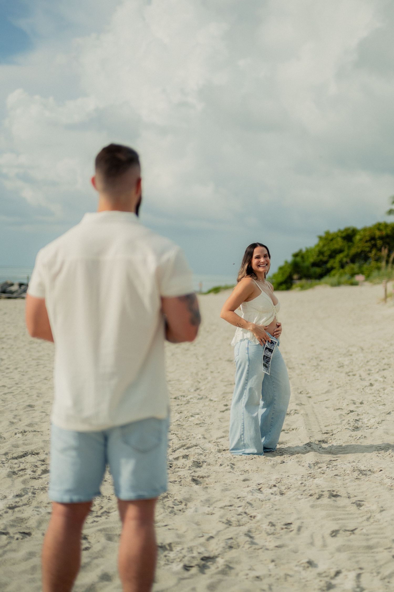 Woman in light blue jeans smiles at a man in a white shirt on a sunny beach.