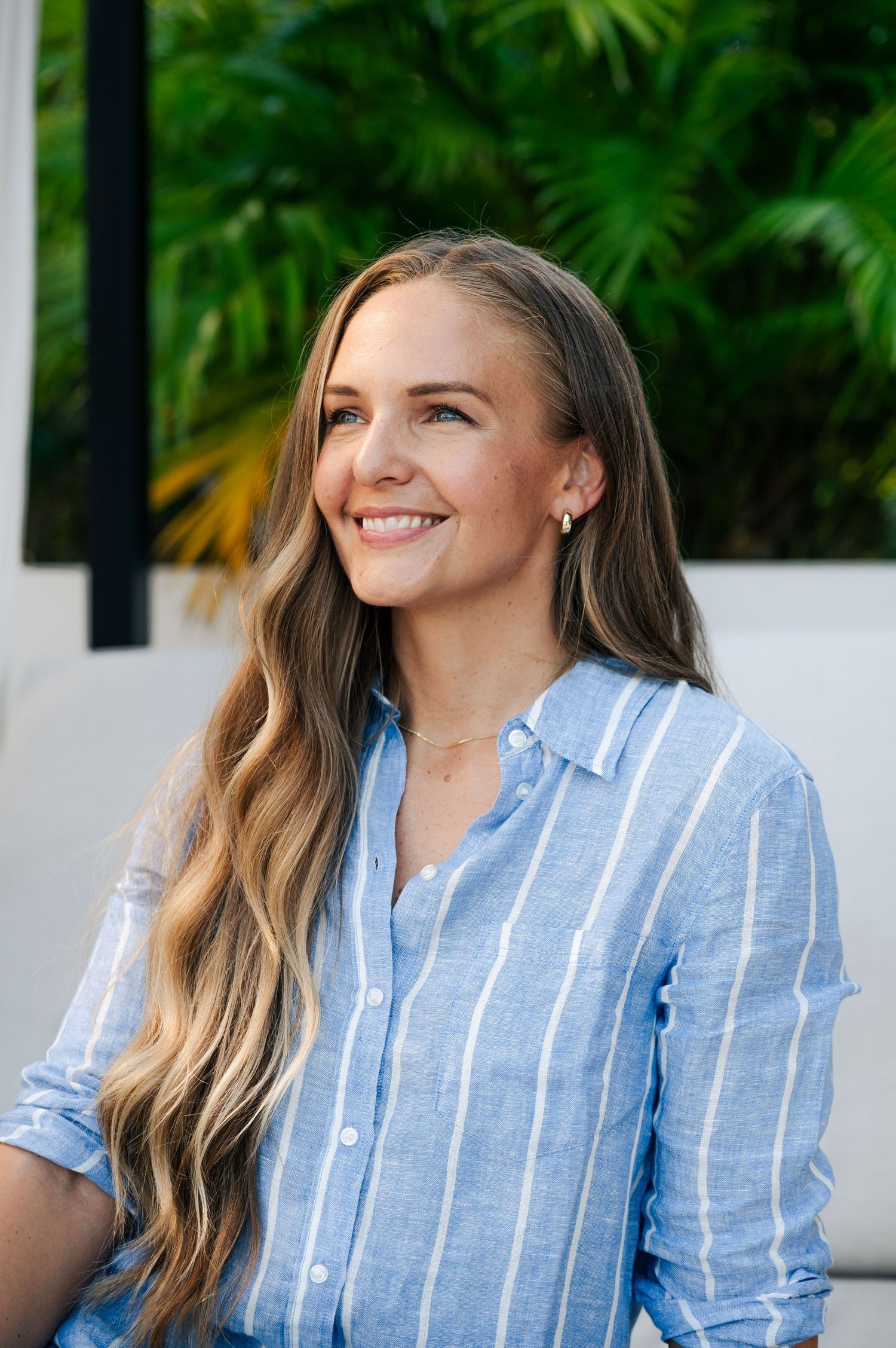 A smiling woman with long wavy blonde hair wears a blue striped linen shirt in front of lush green tropical foliage.