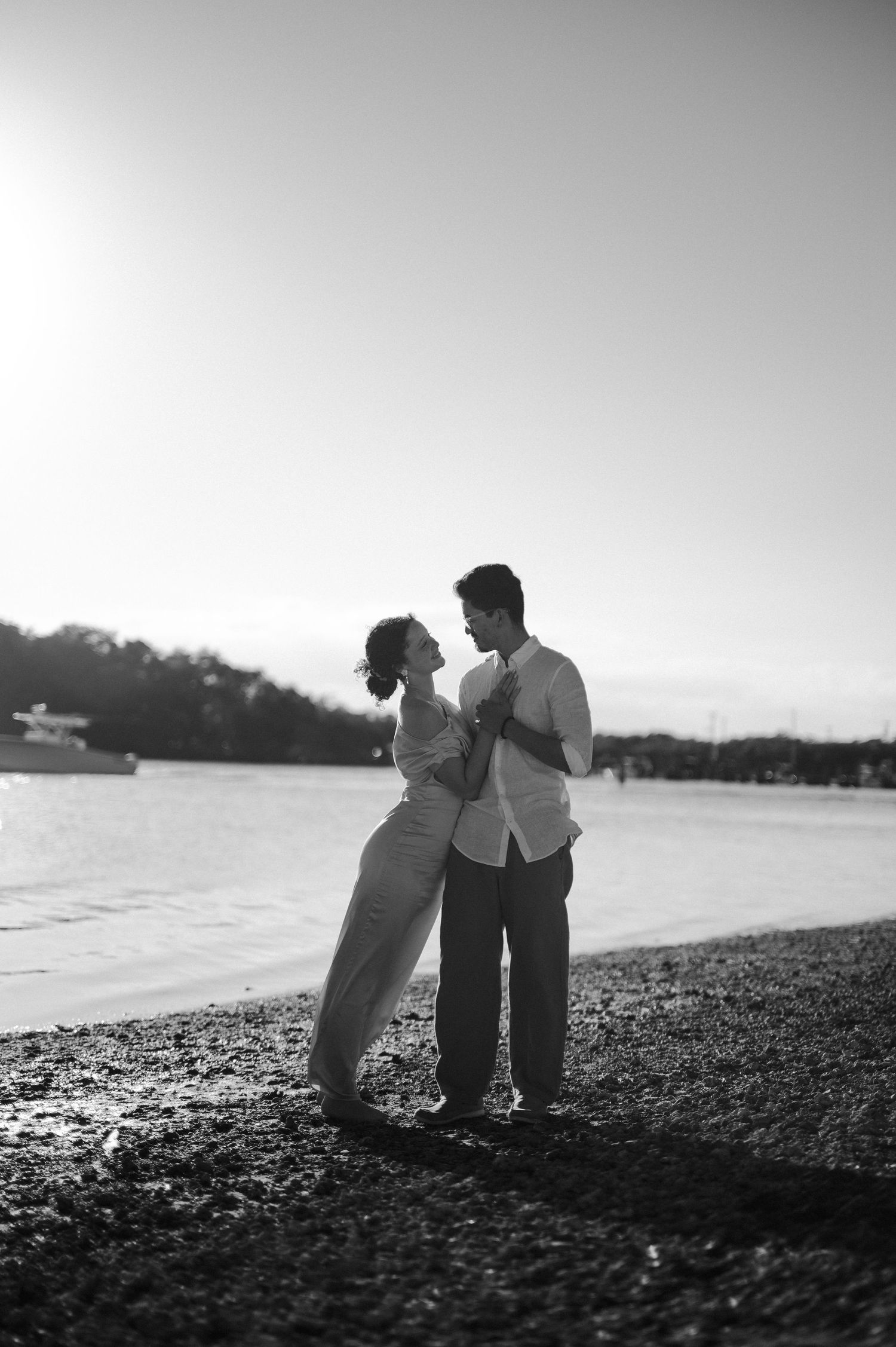 Black and white photo of a couple embracing and gazing at each other on a pebbly shoreline with a boat in the background.