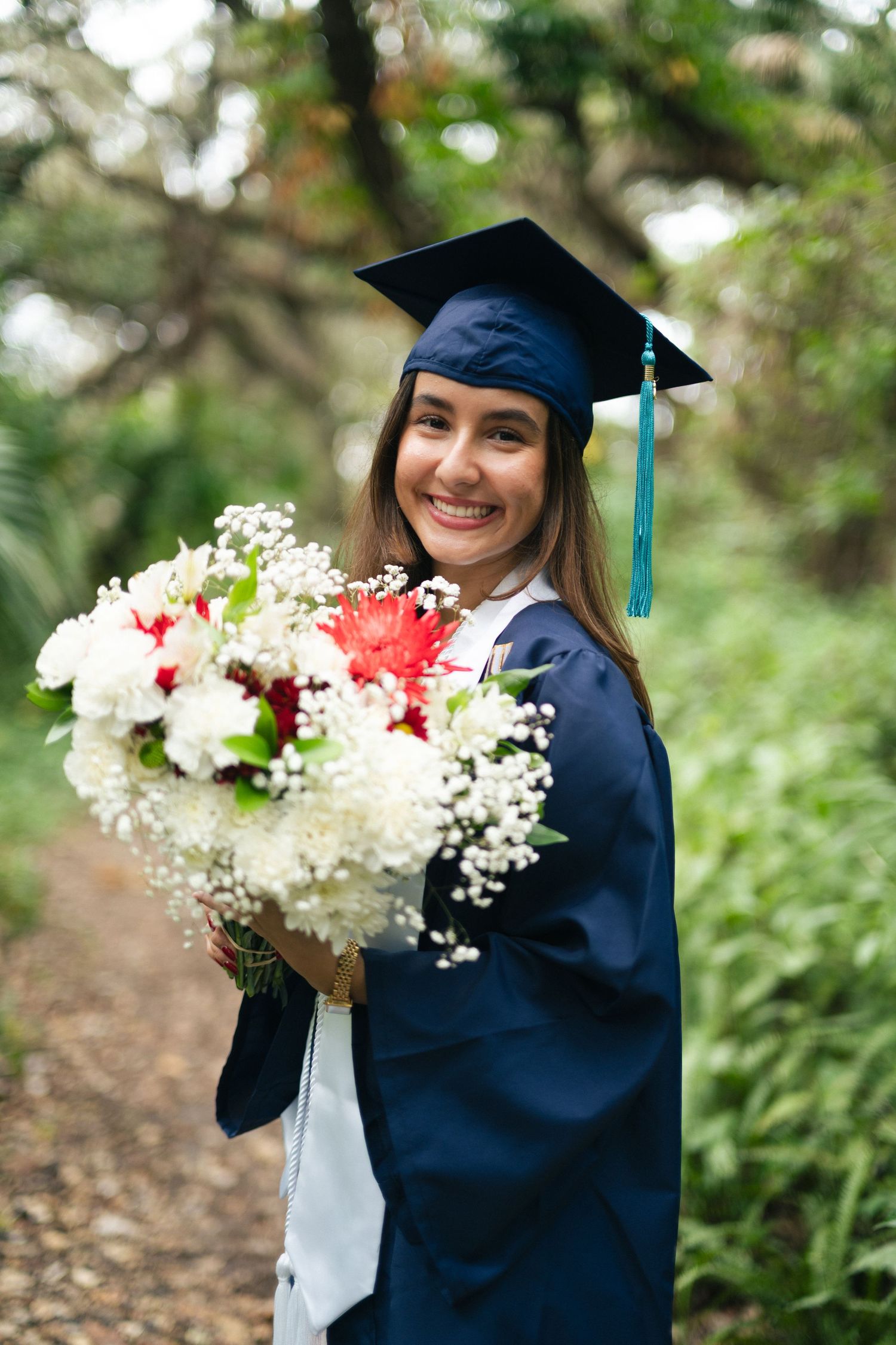 Smiling graduate in a navy cap and gown holds a bouquet of white and red flowers on a wooded trail.