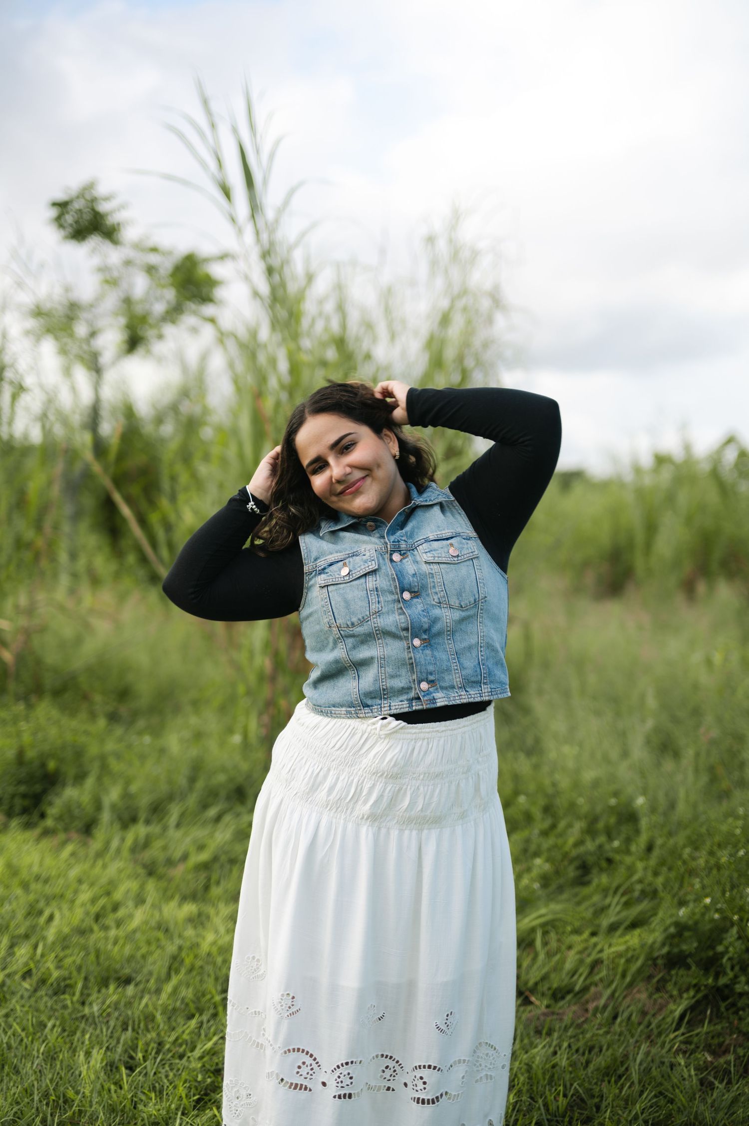 A joyful woman in a denim vest, black long-sleeve top and white lace skirt poses with her hands in her hair among tall green grass.