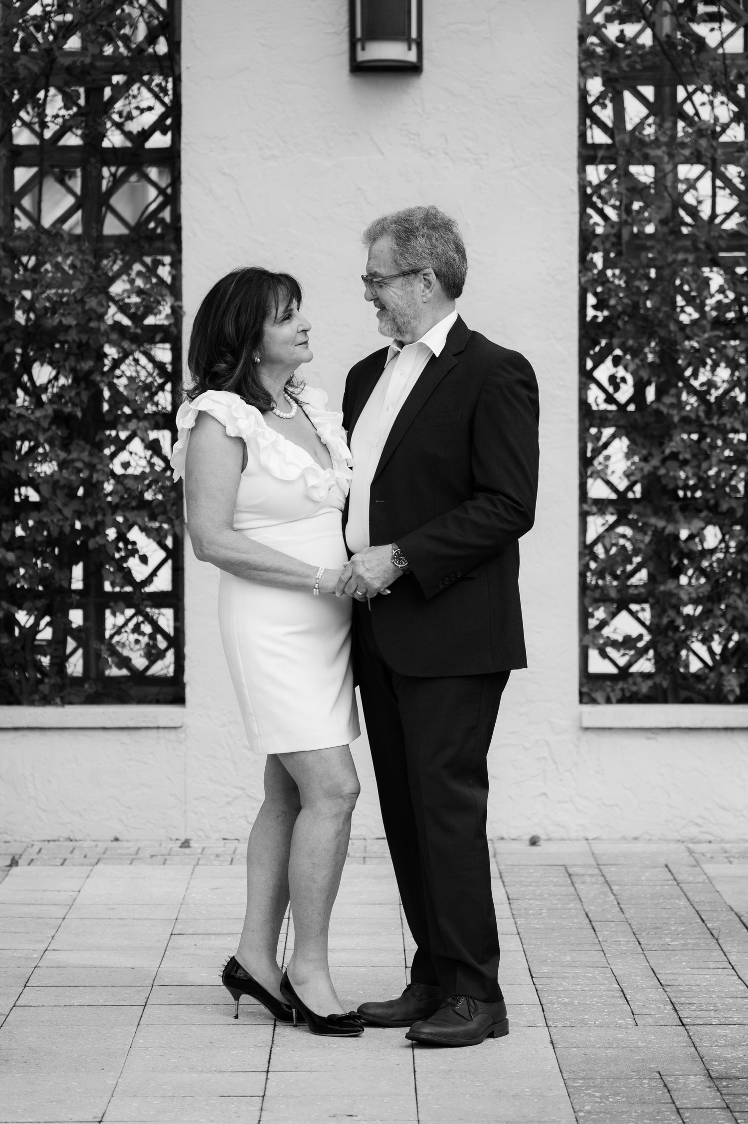 Black and white portrait of a mature couple holding hands and smiling at each other in front of a decorative trellis wall.