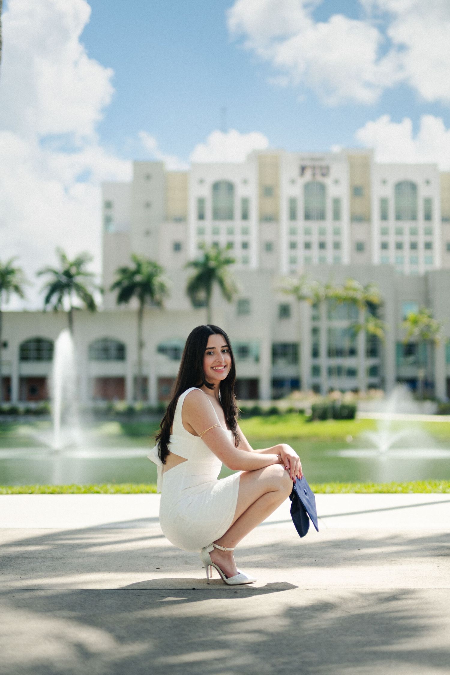 Woman in a white dress crouches smiling by a fountain with a building and palm trees behind her.