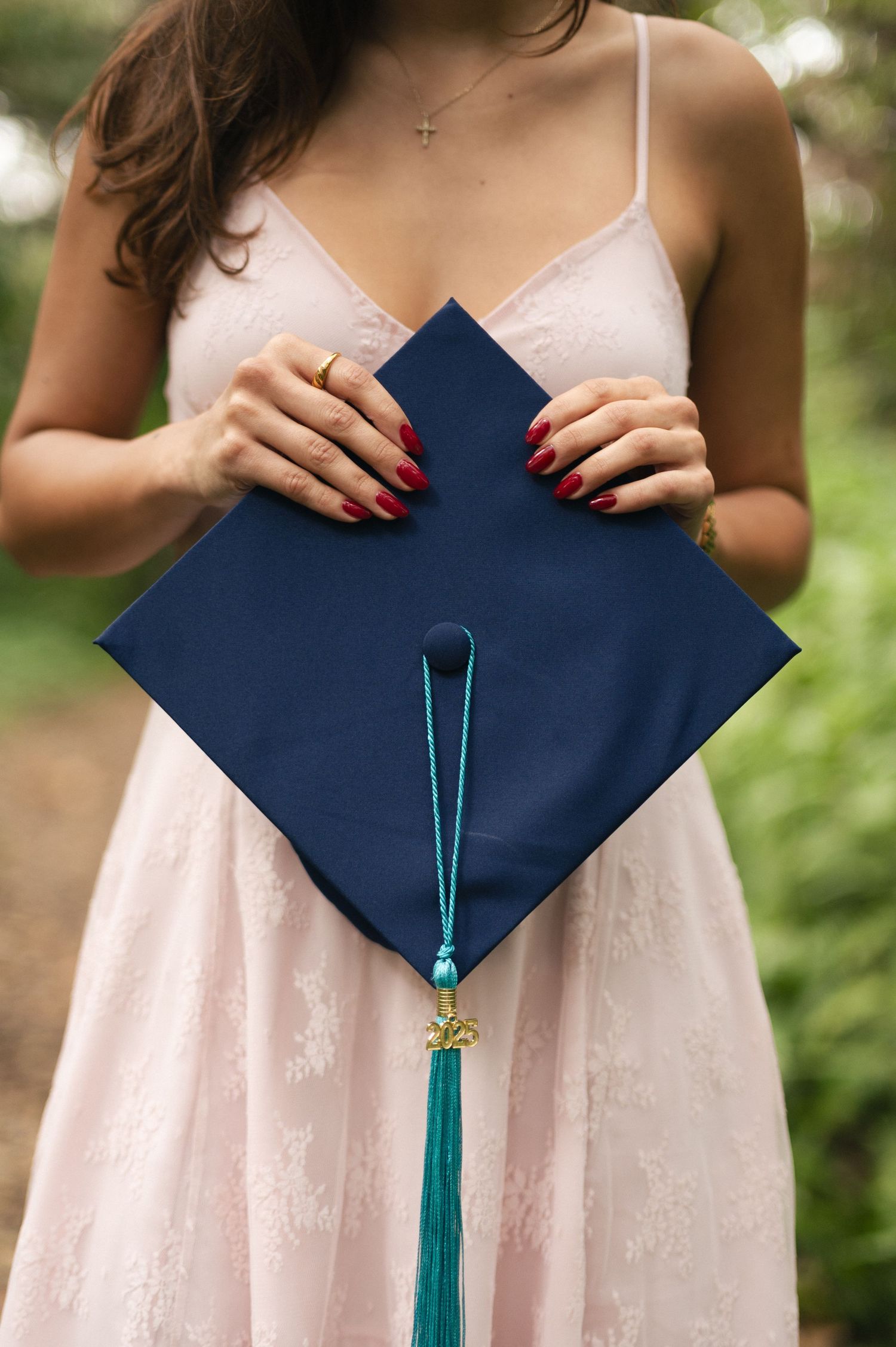 Woman in a pink lace dress holds a navy graduation cap with a teal tassel, showing red painted nails.