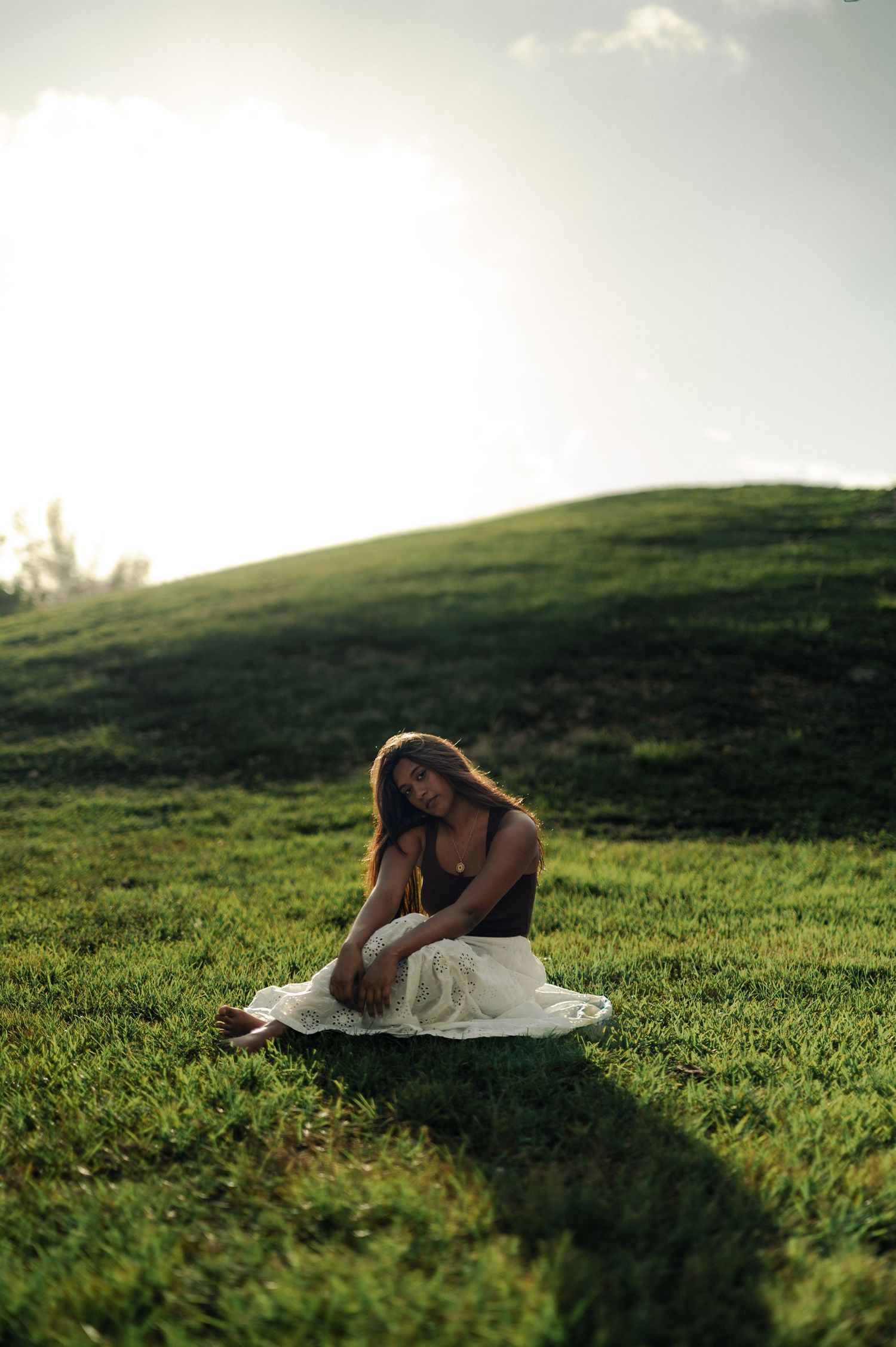 A woman with long hair sits on a green grassy hill in a white skirt, glowing in warm golden backlight.