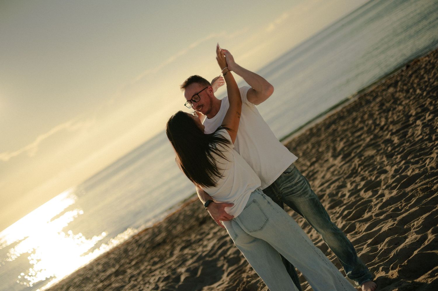 Couple dances playfully on the sandy beach at sunset, the man twirling the woman as golden light reflects on the ocean.