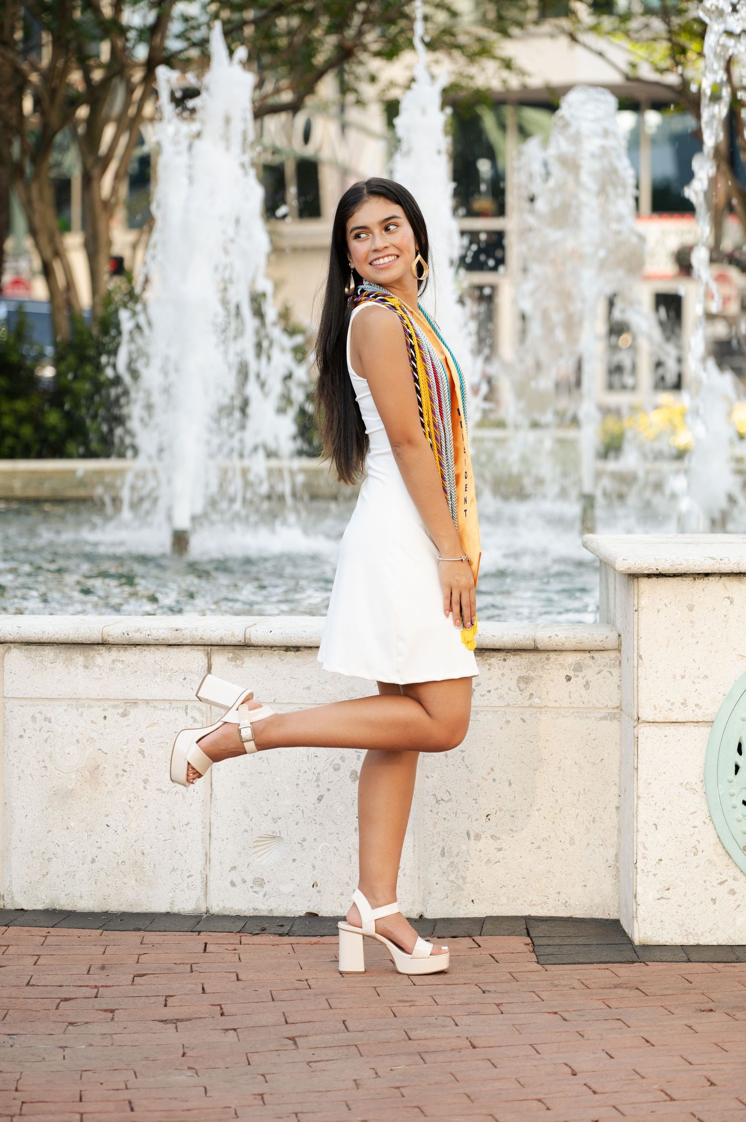 Graduate in a white dress and graduation posing beside a fountain at FIU in Miami.