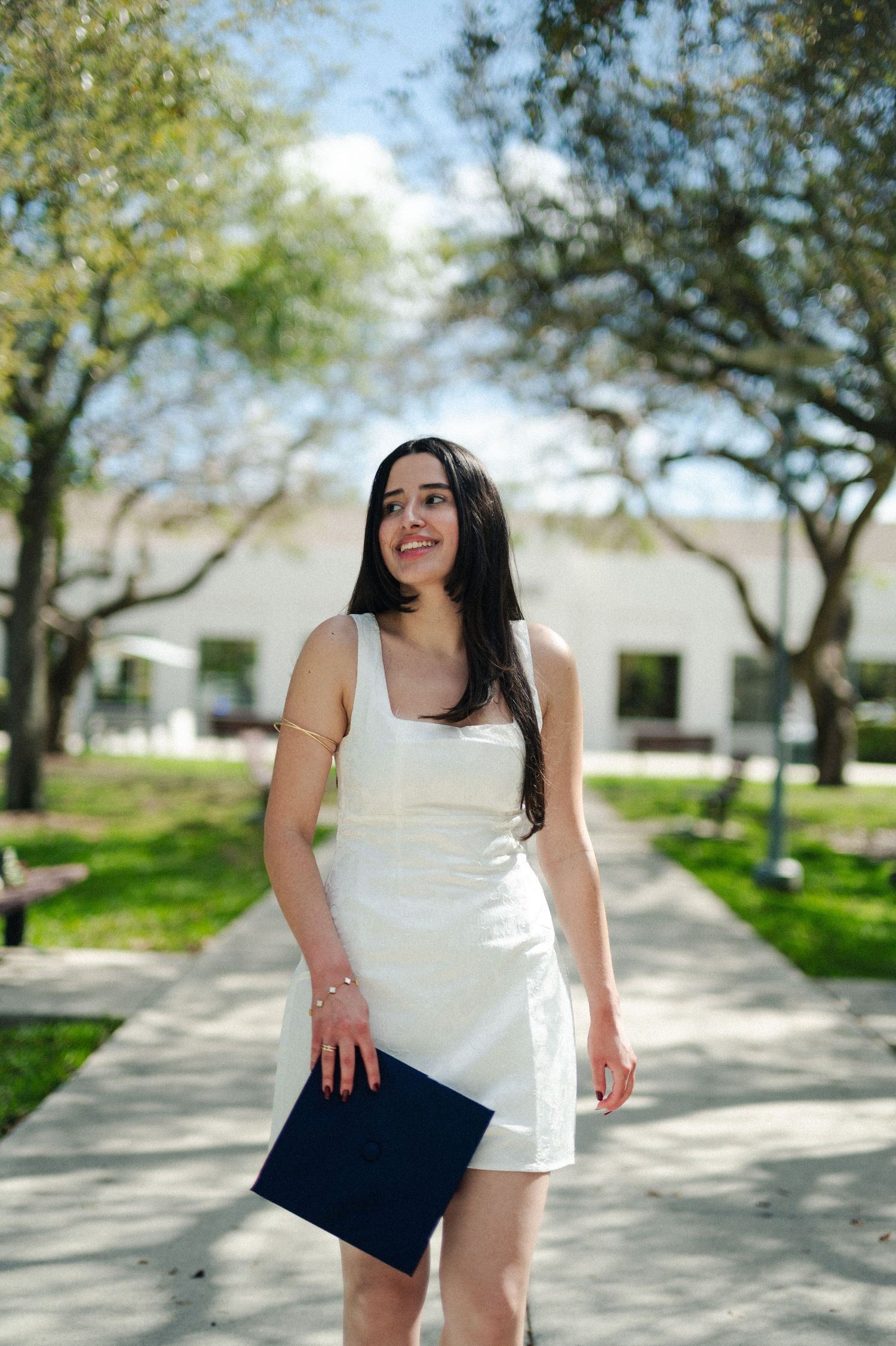 Woman in a white dress smiles on a tree-shaded campus path while holding a navy graduation cap.