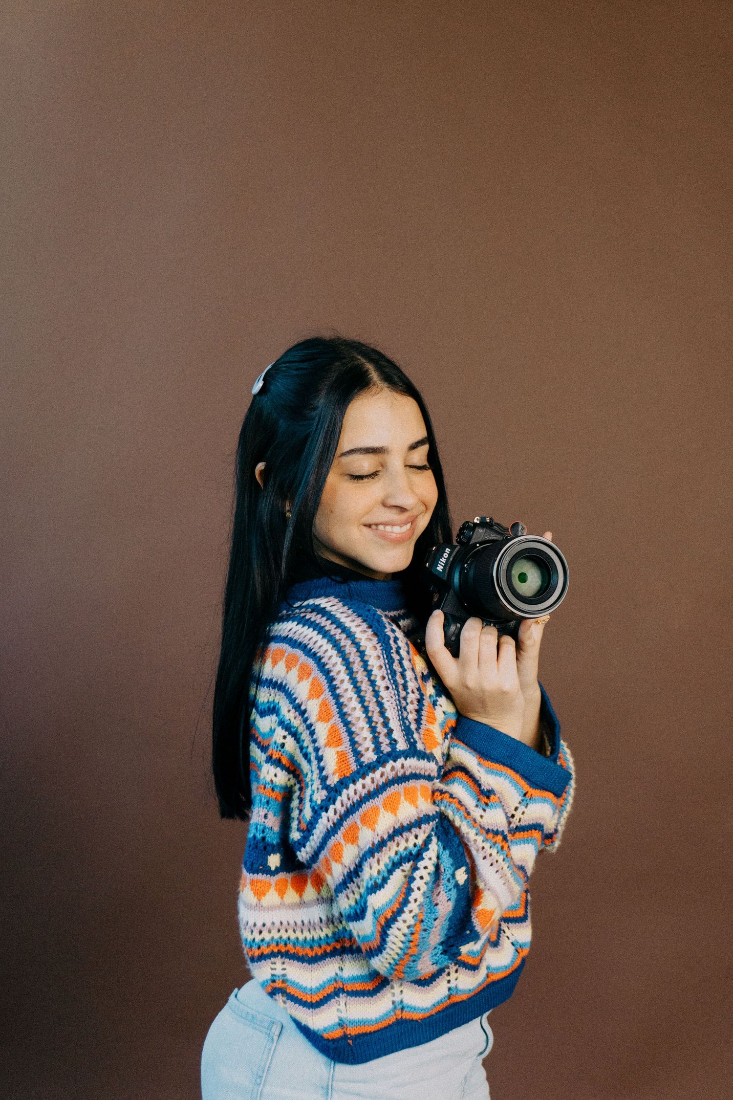 Smiling woman with long dark hair in a colorful patterned sweater holding a camera against a brown backdrop.