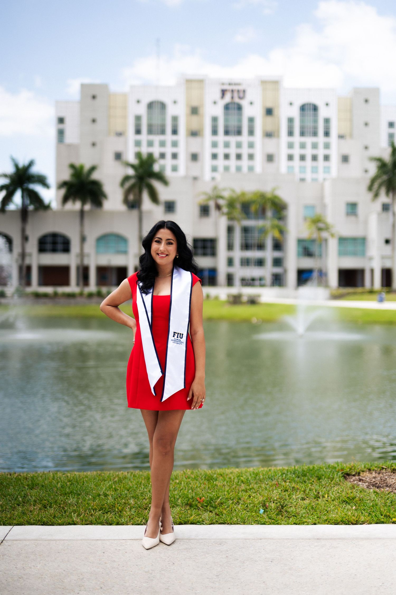 Graduate in a red dress and white university sash stands before a pond and a large campus building with palm trees.