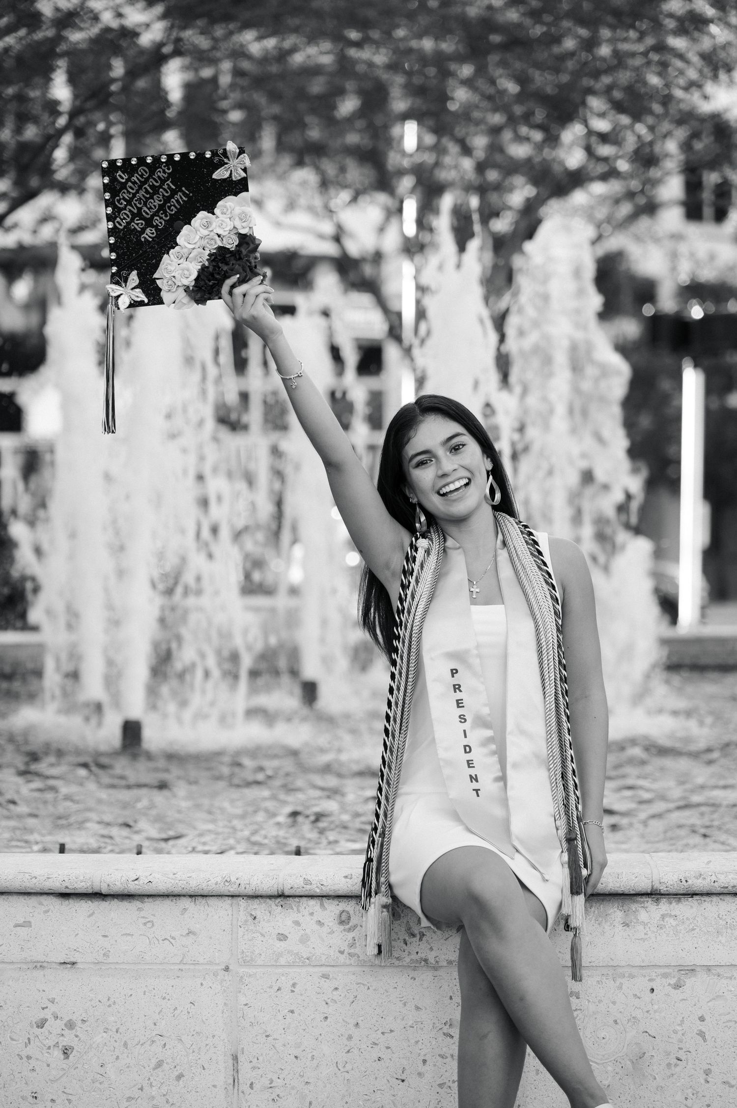 Black and white photo of a joyful graduate in a white dress raising her decorated cap beside a fountain.