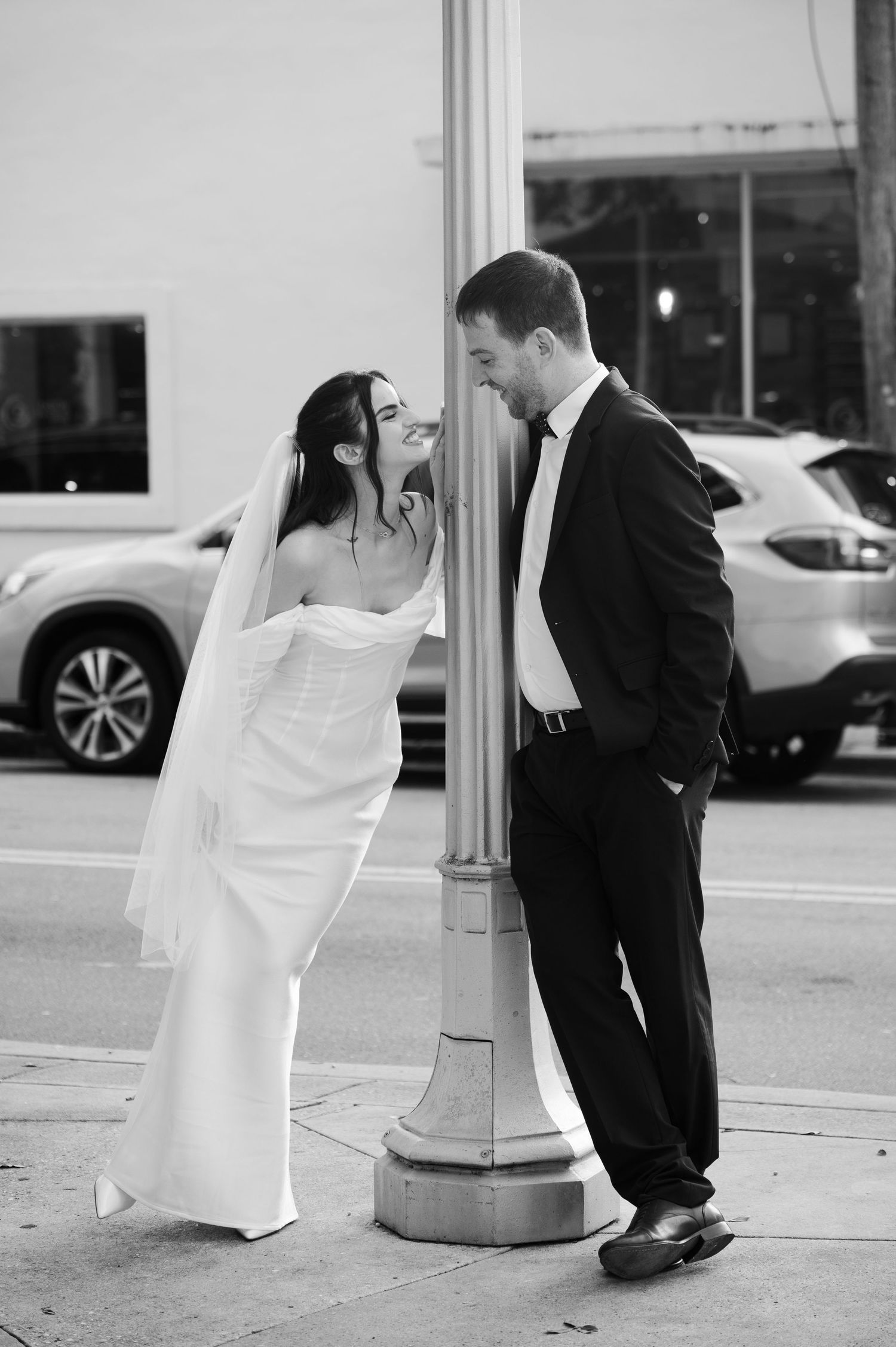 A black and white photo of a bride in a white gown and veil leaning toward a groom in a suit against a street lamppost.