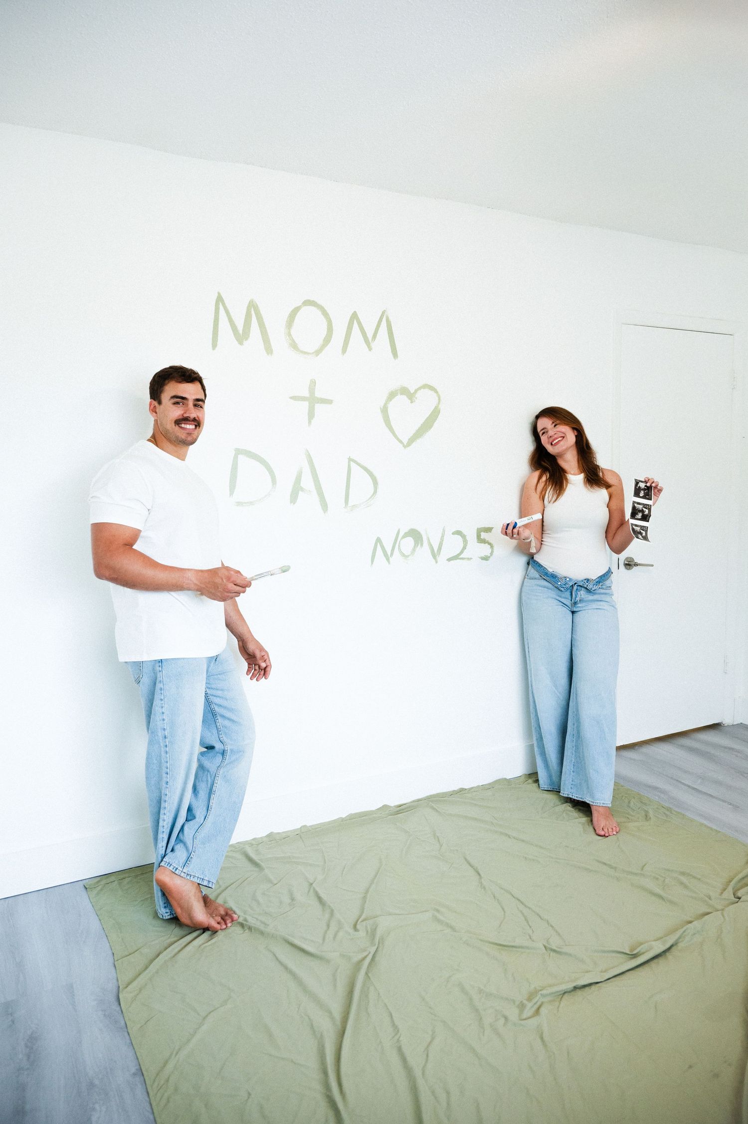Smiling couple stands before a white wall with a Mom plus Dad pregnancy announcement and due date painted in green.