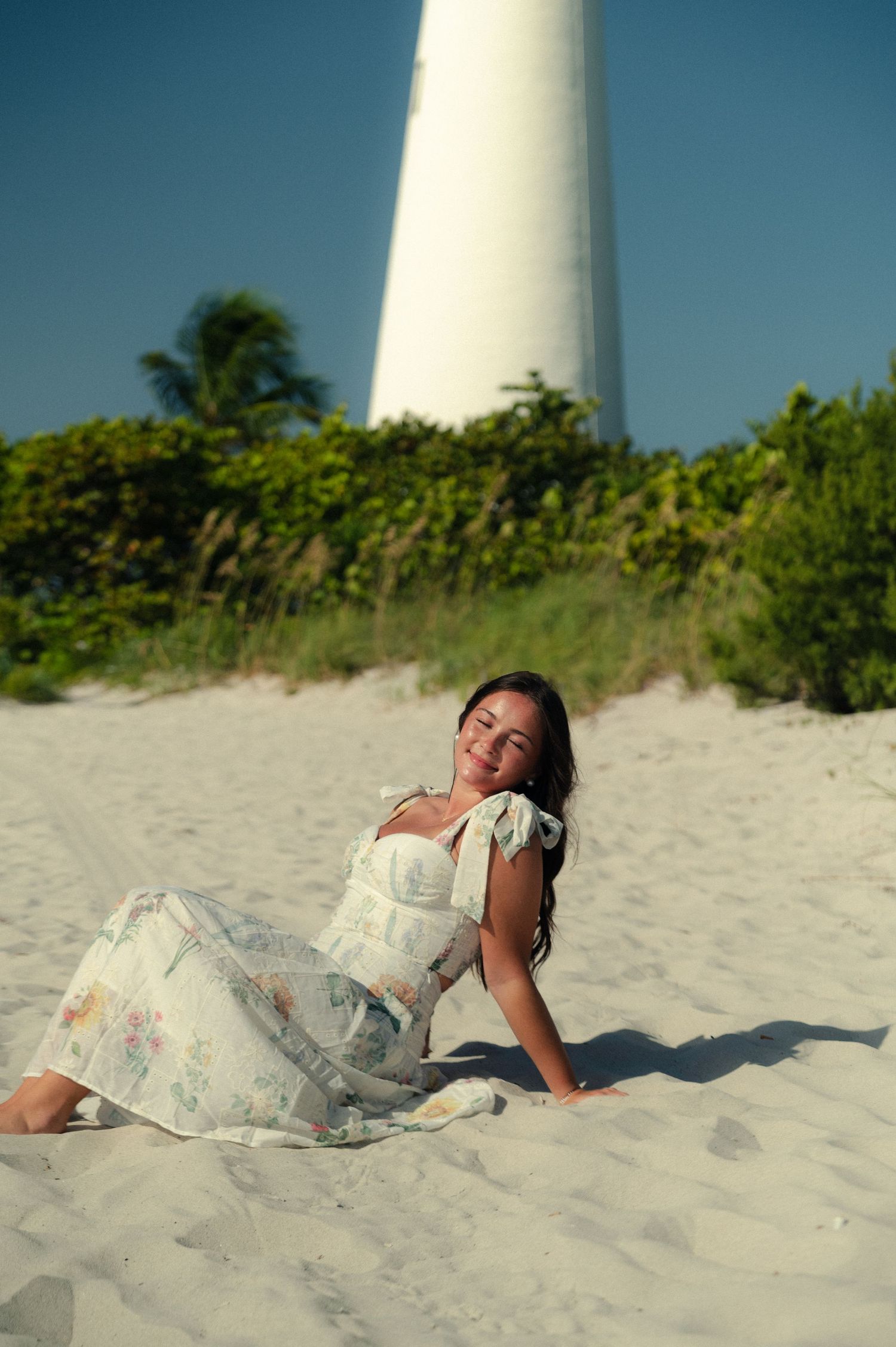 Woman in a floral dress relaxes on white sand with eyes closed in front of a white lighthouse and blue sky.